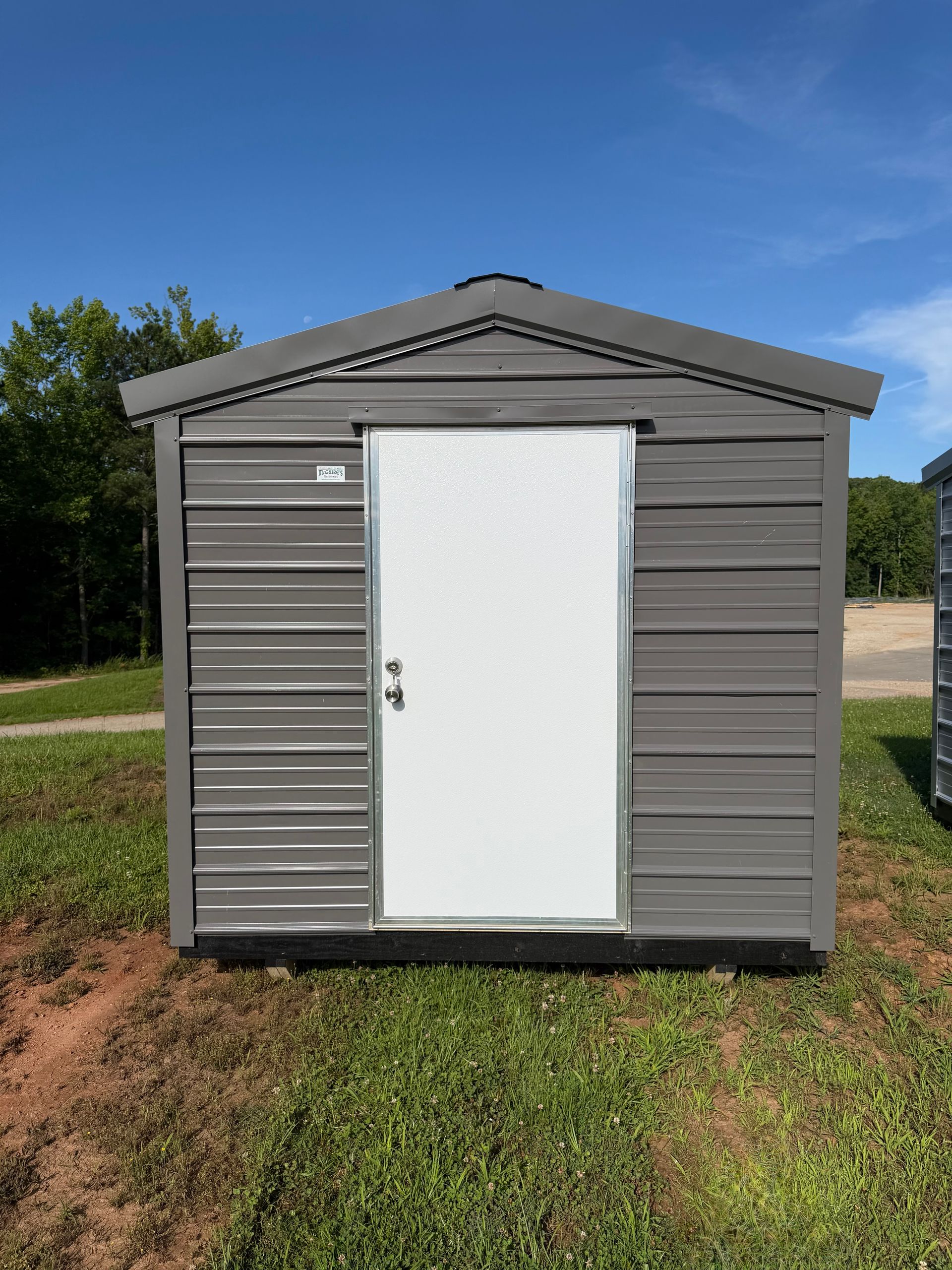 Small gray shed with a white door on a grassy lot under a blue sky