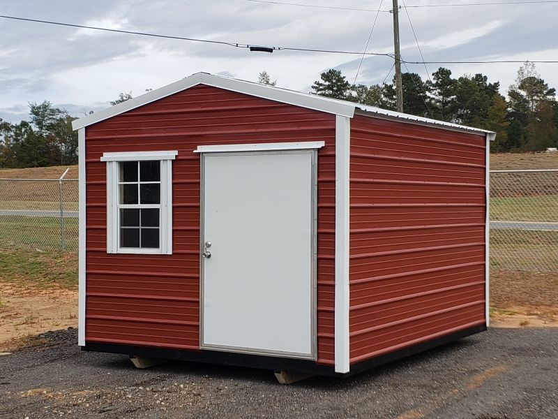 Red metal storage shed with white door, window, and trim, set outdoors on a gray surface.