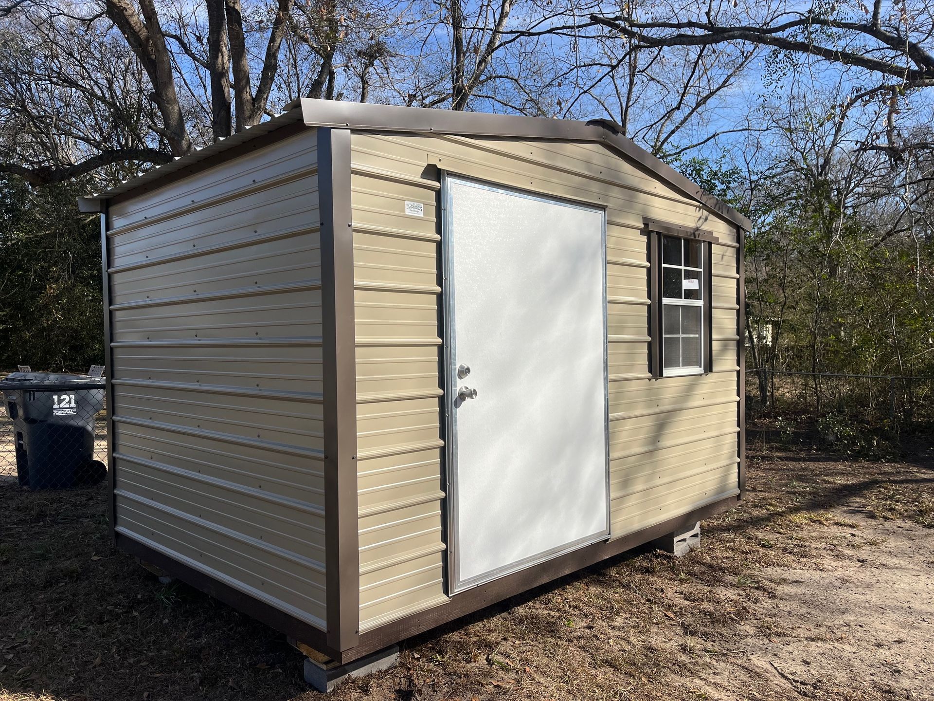 Tan metal storage shed with a white door and small window, set outside on a sunny day.