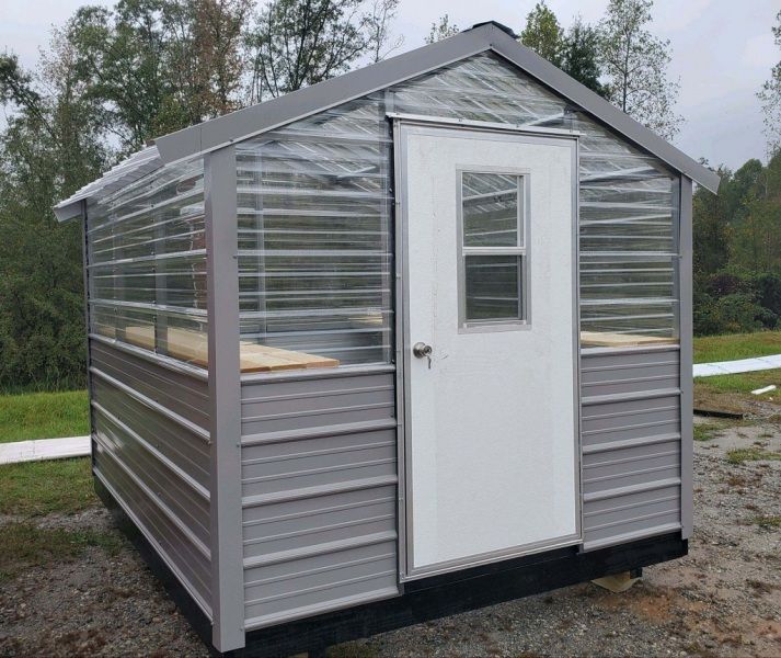 Gray and clear-walled greenhouse with white door, set outdoors under overcast sky.