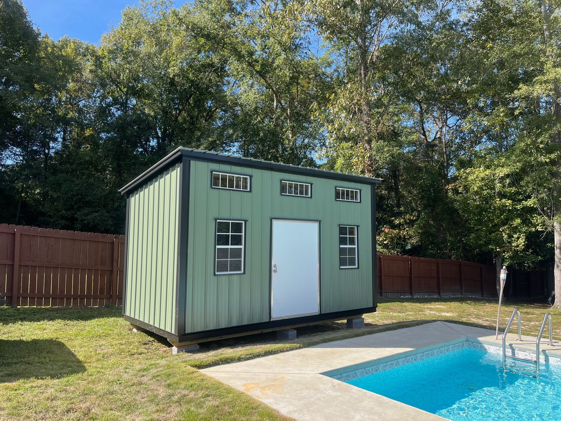 Green shed with white door and windows, near a pool, set against a brown fence and trees.