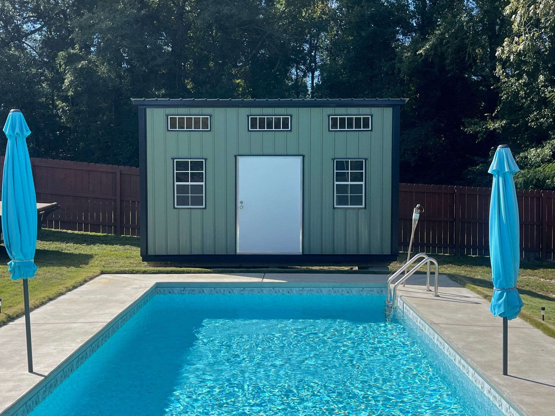 Poolside shed with teal exterior, white door, and small windows; two blue umbrellas on the sides.