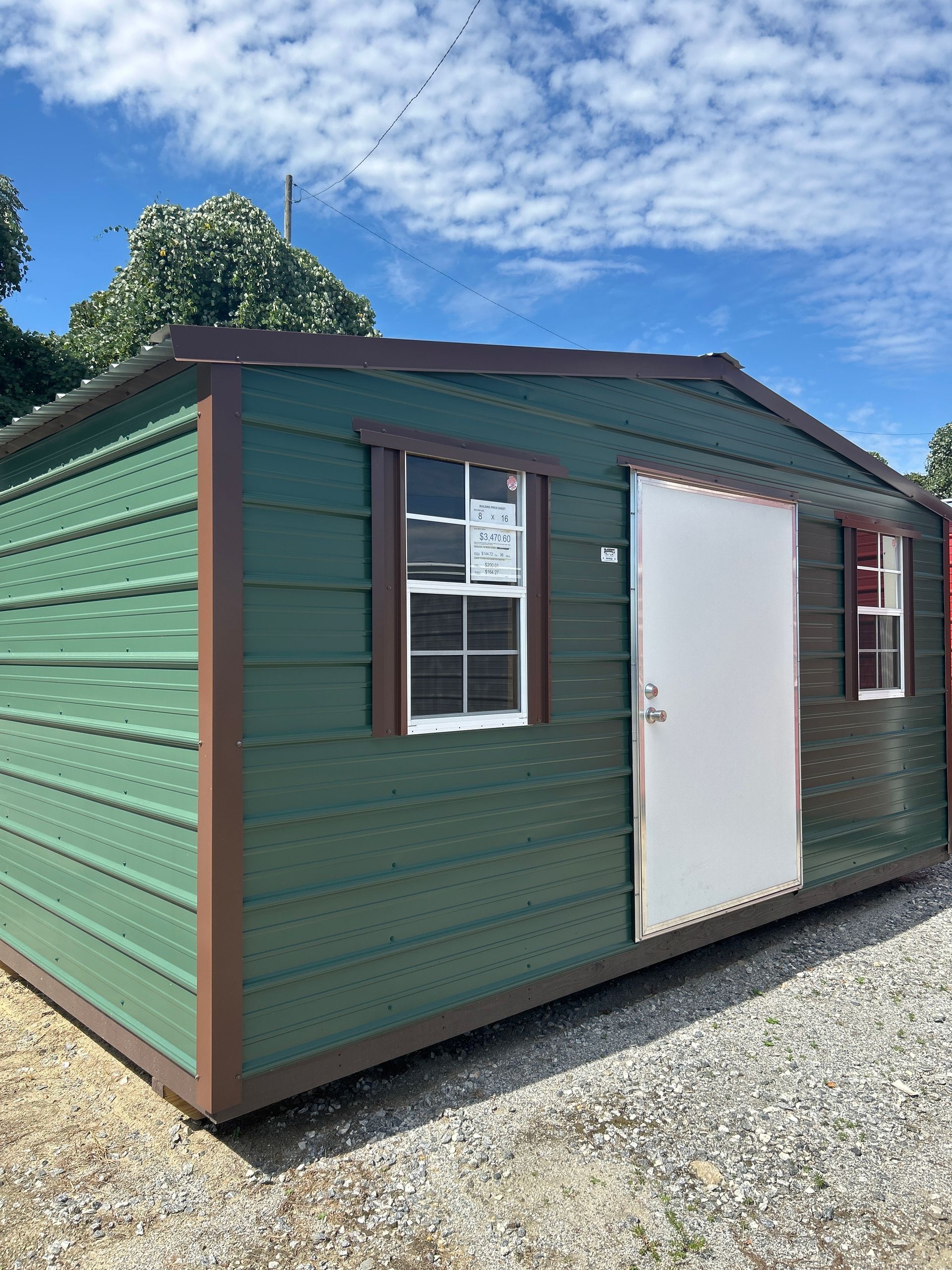 Green storage shed with brown trim, white door, and two windows, set outdoors under a blue sky.
