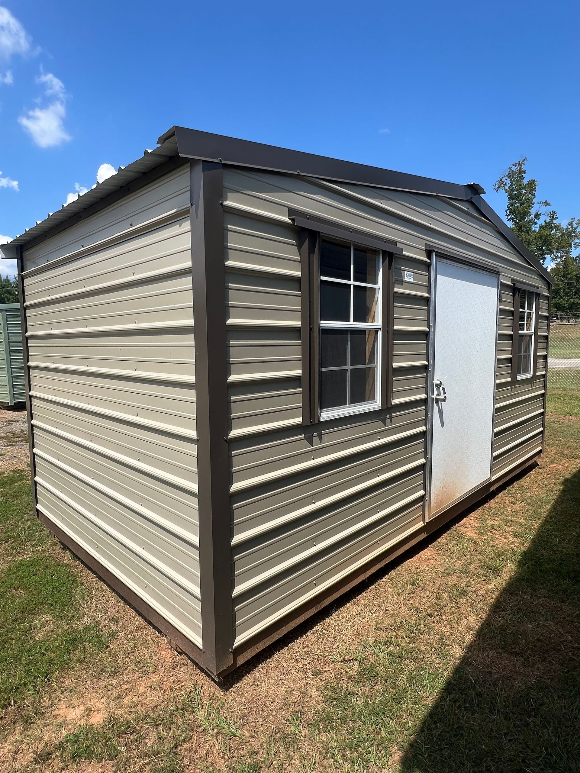 Tan and brown metal shed with a black roof, two windows, and a white door, on grassy ground.