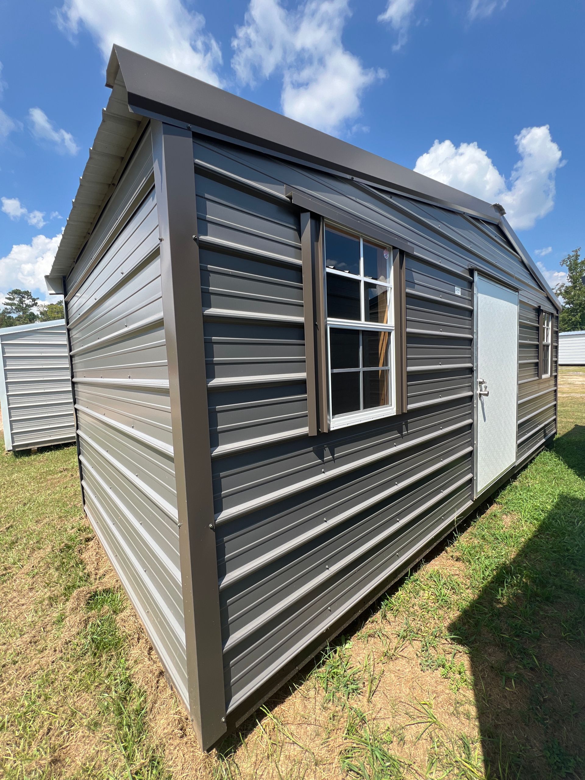 Gray metal shed with a brown roof, window, and door on a grassy field under a blue sky.