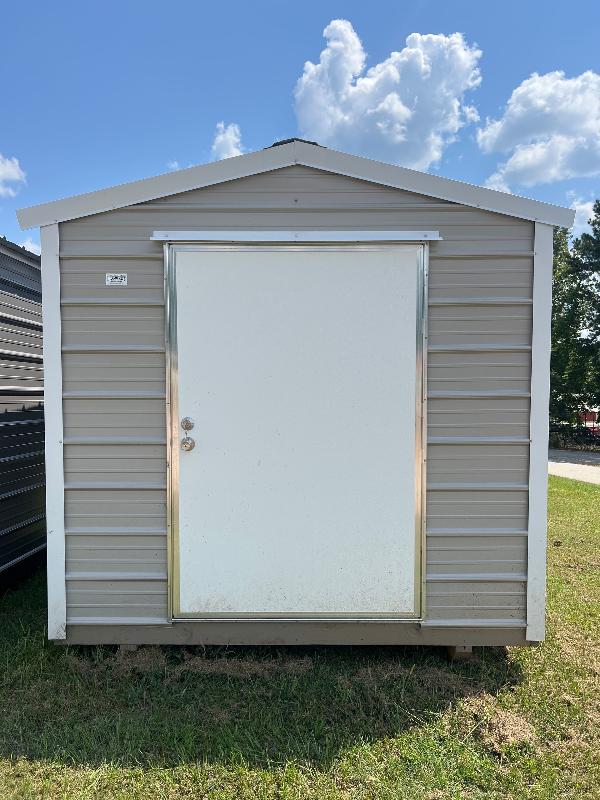 Gray shed with white door, set on green grass, against a blue sky with clouds.