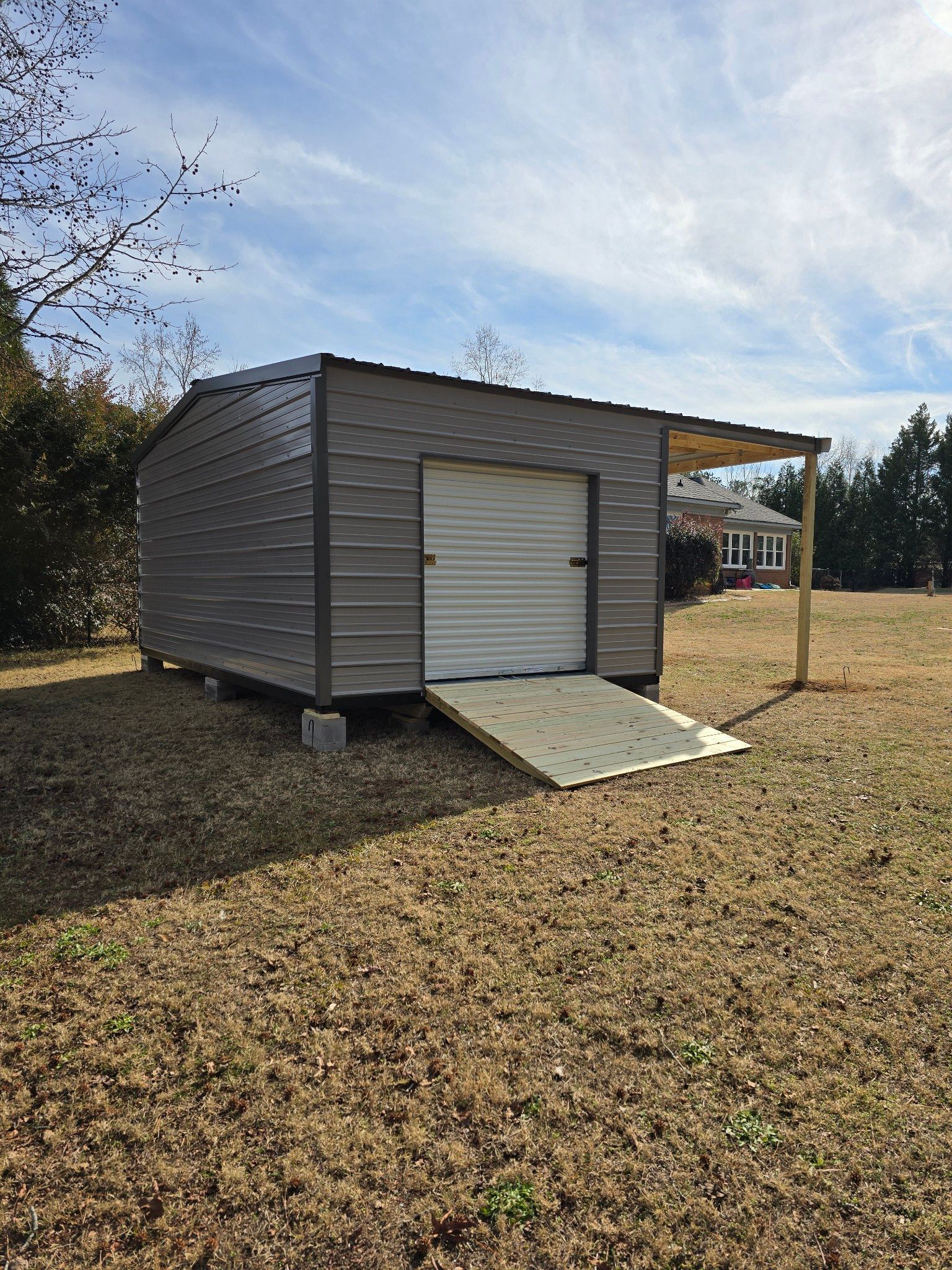 A gray metal shed with a ramp and small awning in a grassy yard under a blue sky.