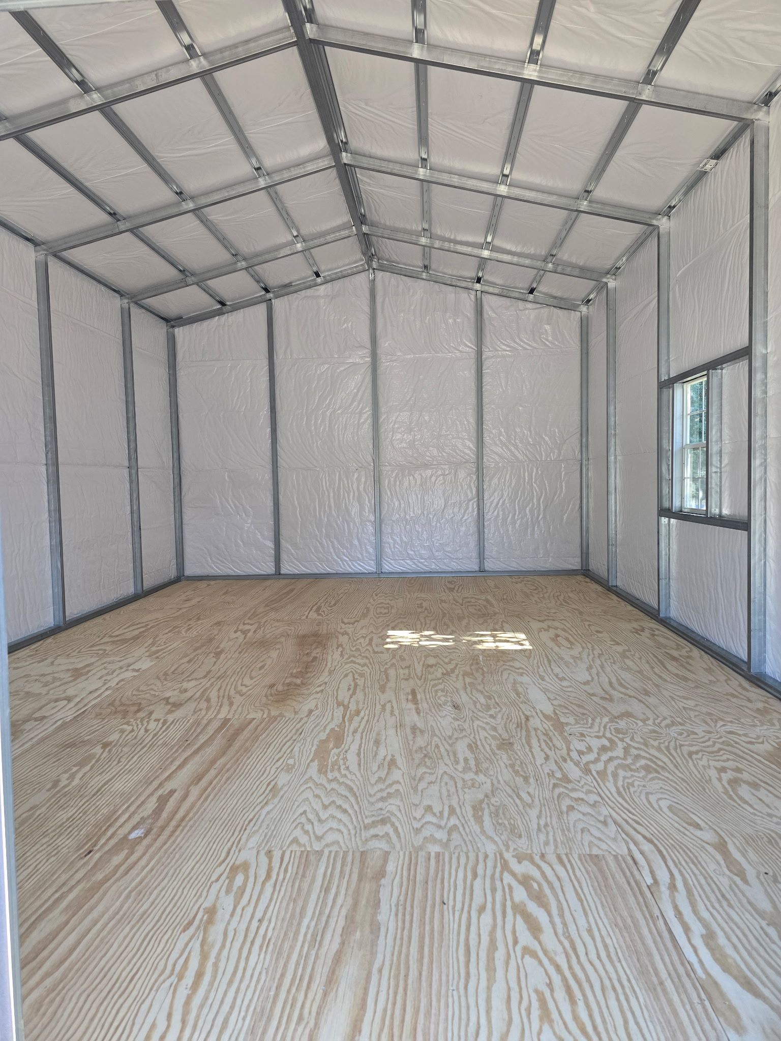 Interior of an empty metal-framed storage shed with plywood floor, white walls, and a small window.