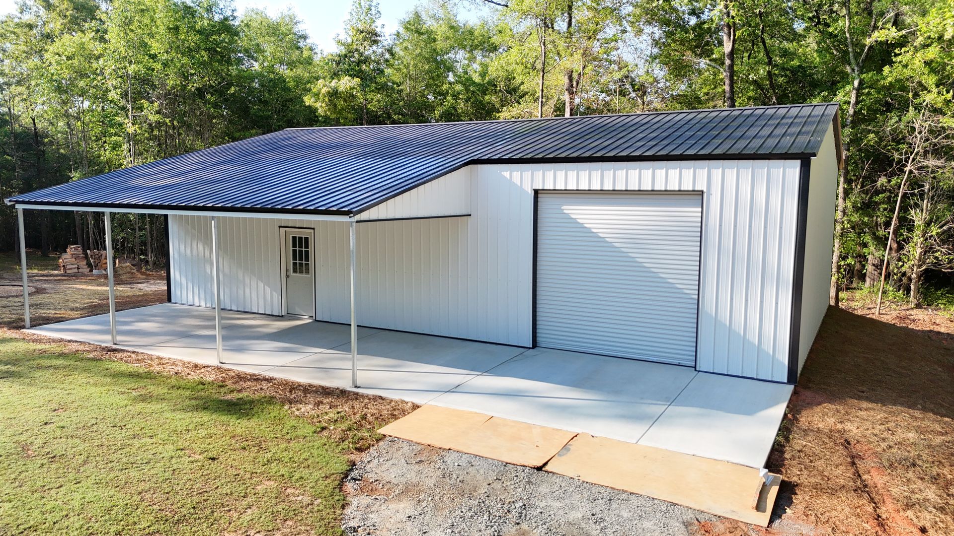 White metal building with a black roof and a concrete porch in a grassy area with trees. It has a garage door and a door.
