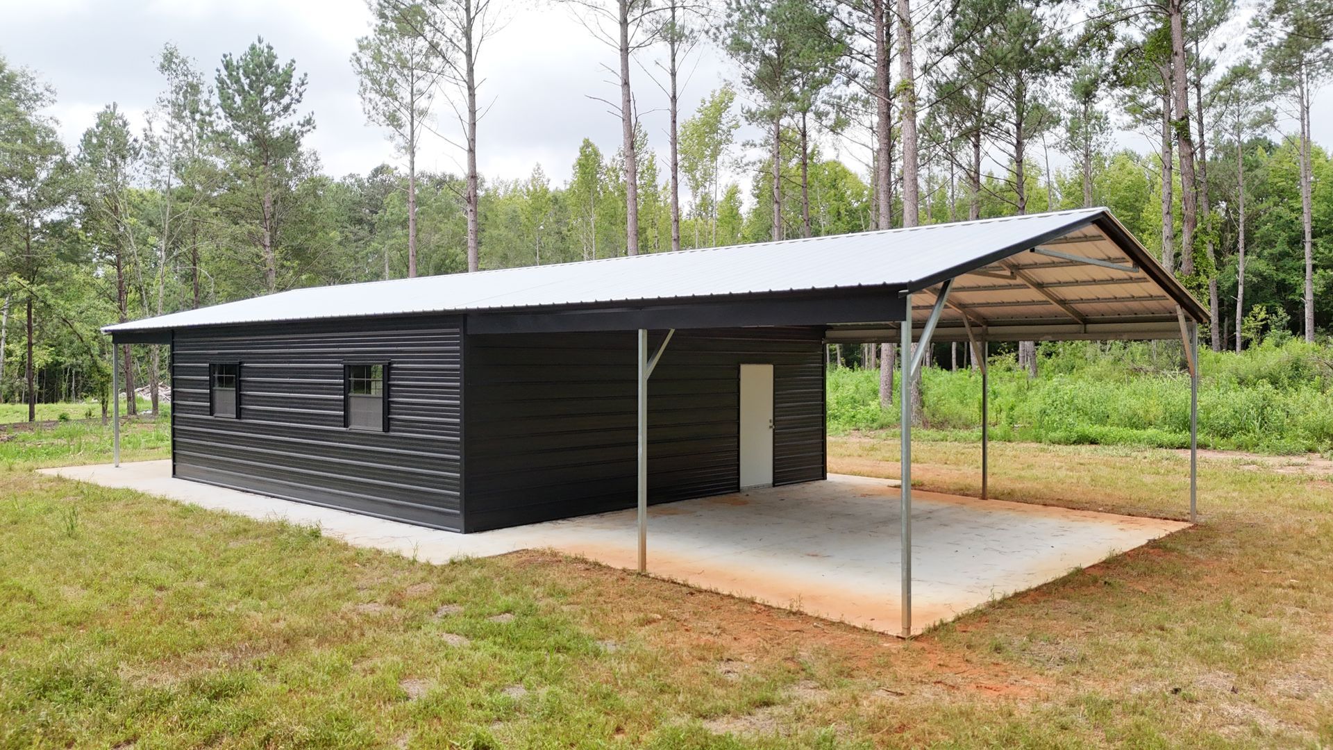 Black metal building with a carport, set in a grassy area with trees in the background.