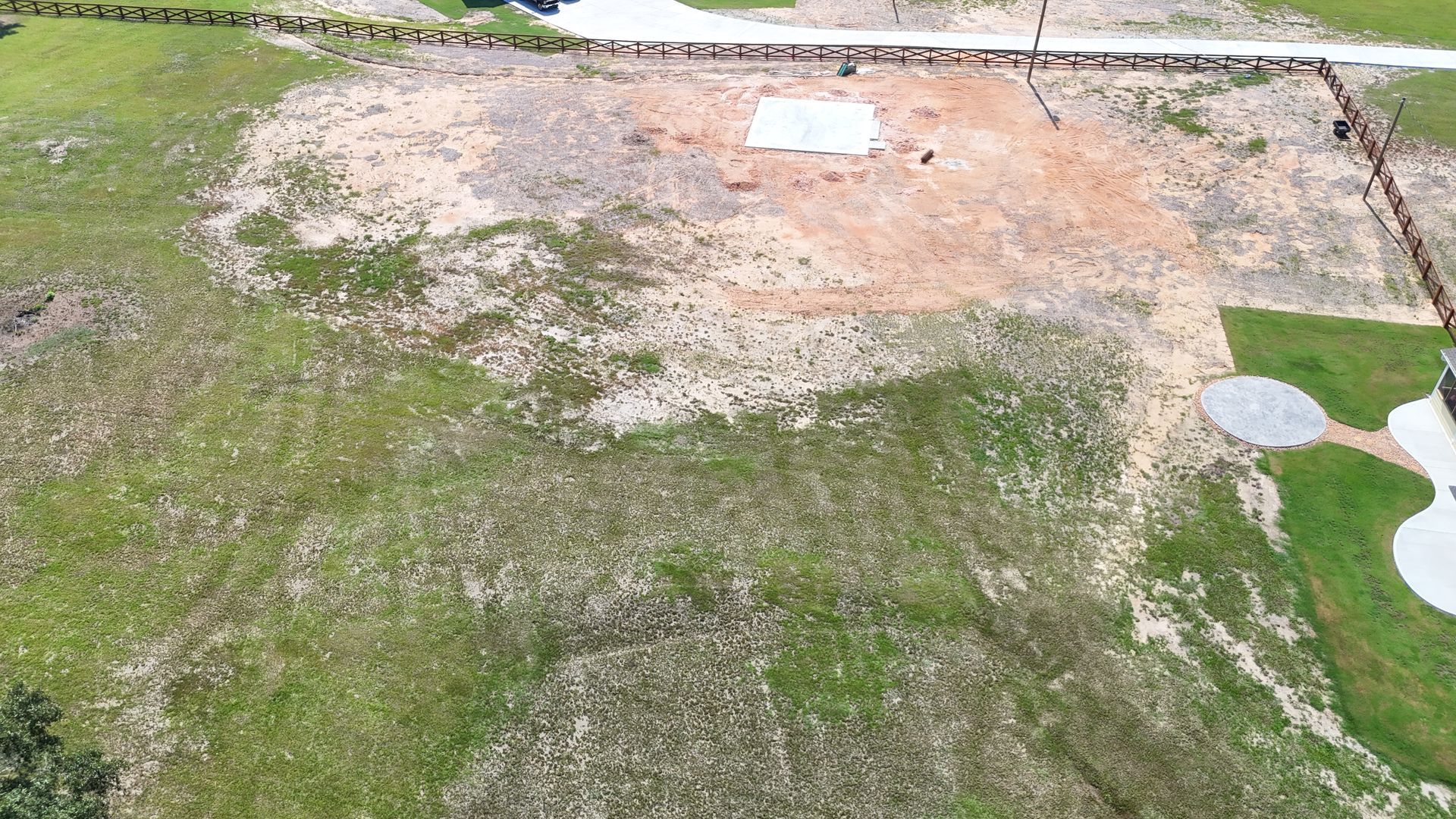 Aerial view of a cleared construction site with a concrete foundation pad, surrounded by grassy areas and a fence.