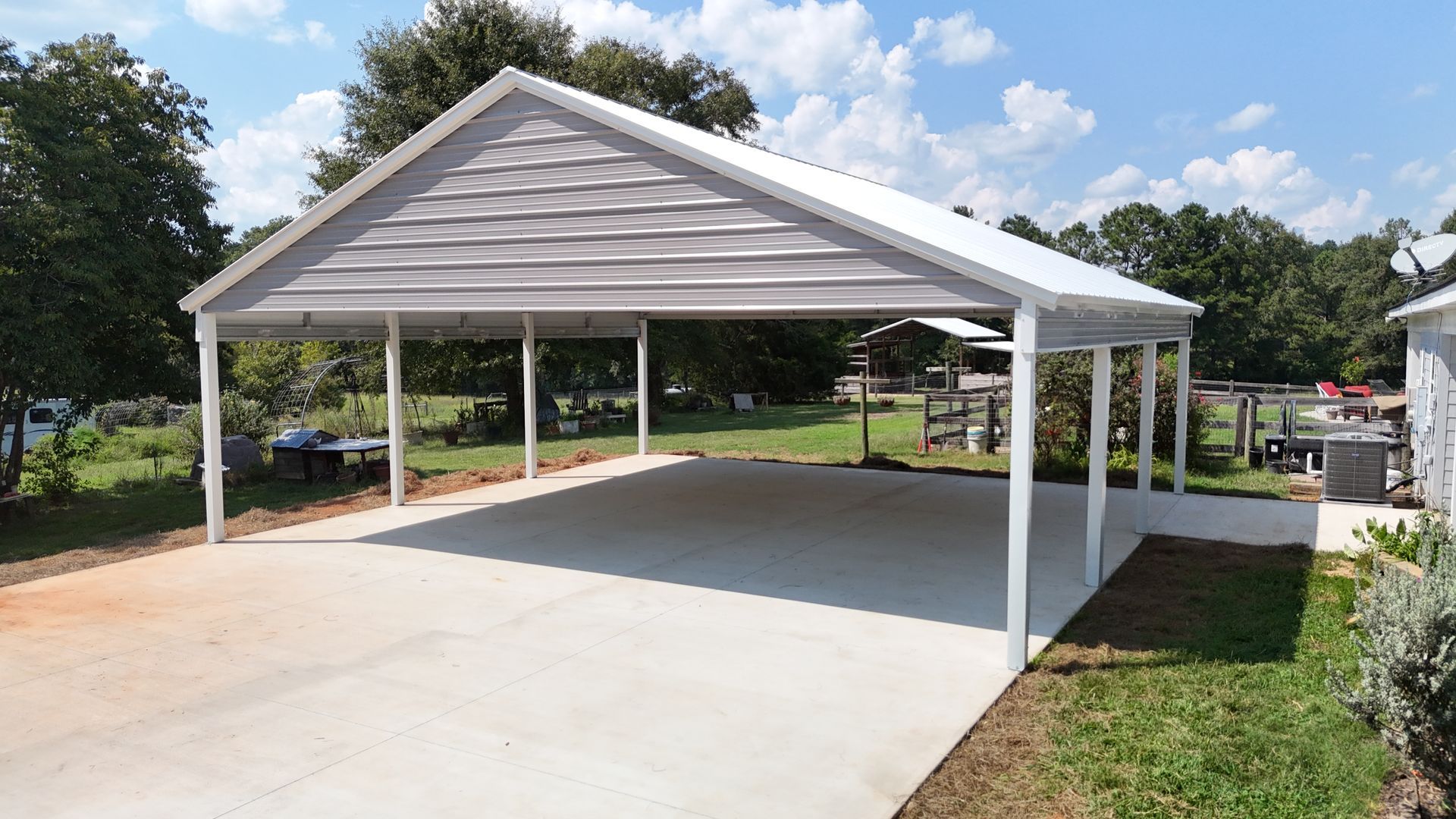 A carport with a gray roof and white supports sits on a concrete pad.