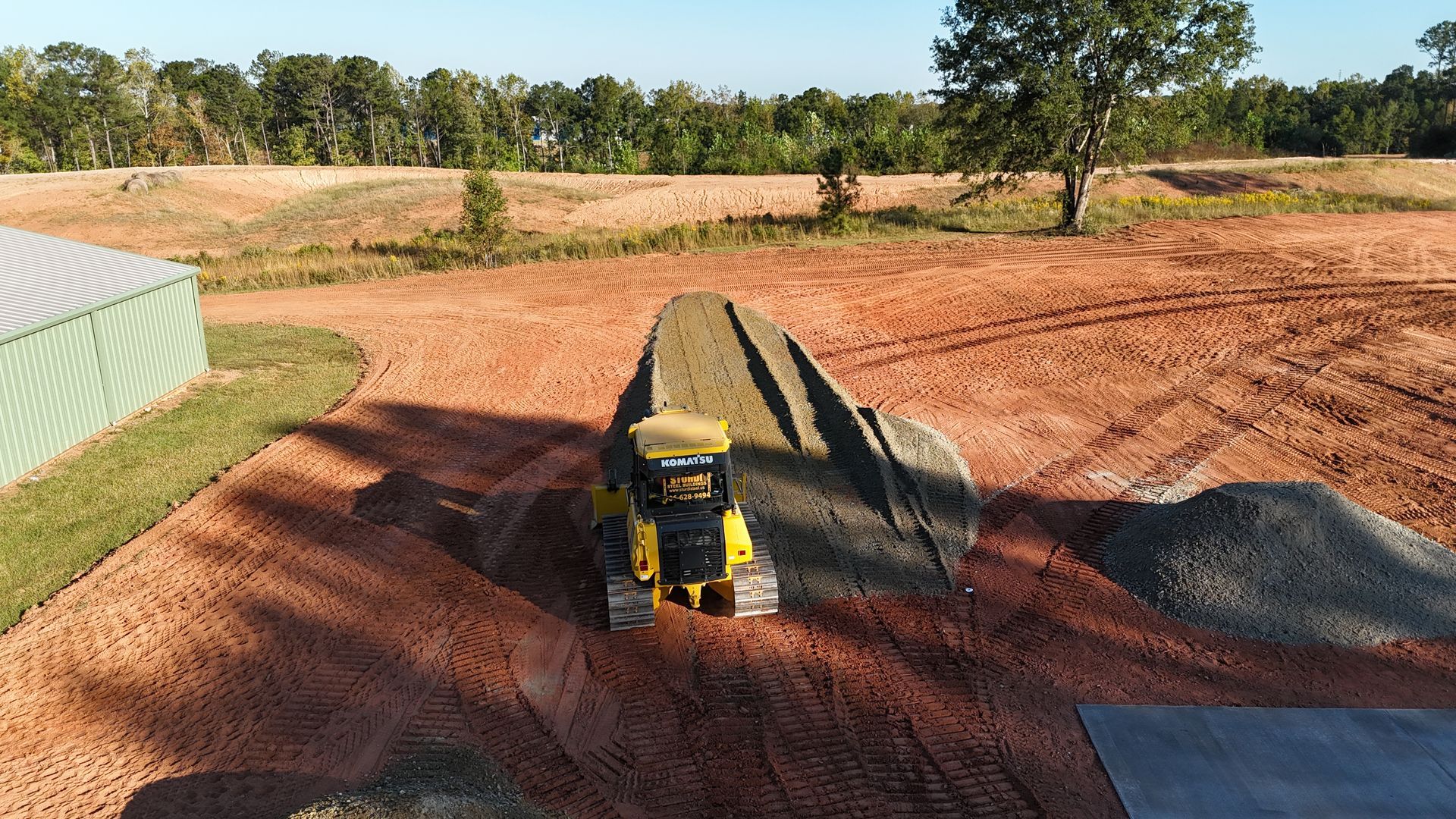 Yellow tractor grading a gravel pile on a reddish dirt field, near a green metal building and trees.