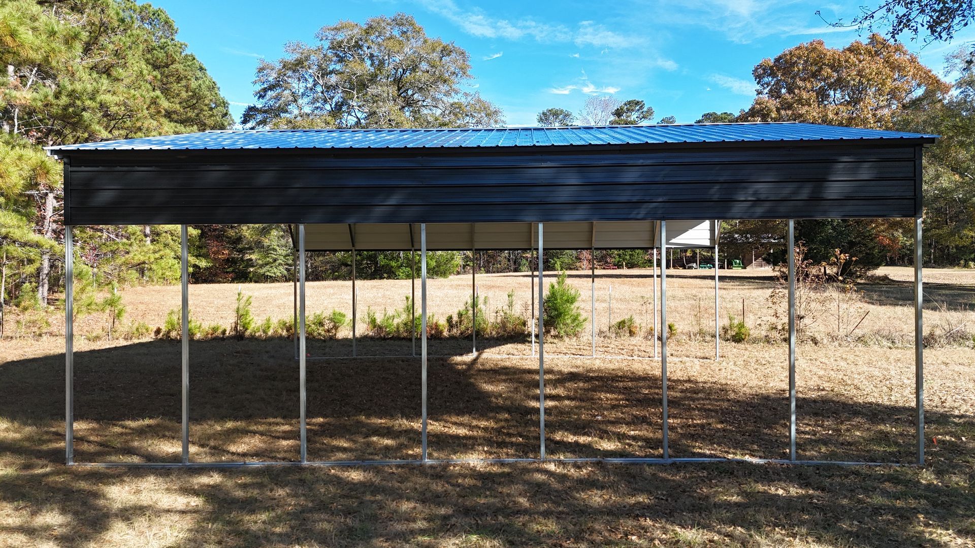 Black carport with metal roof and silver support beams, set in a grassy field with trees.