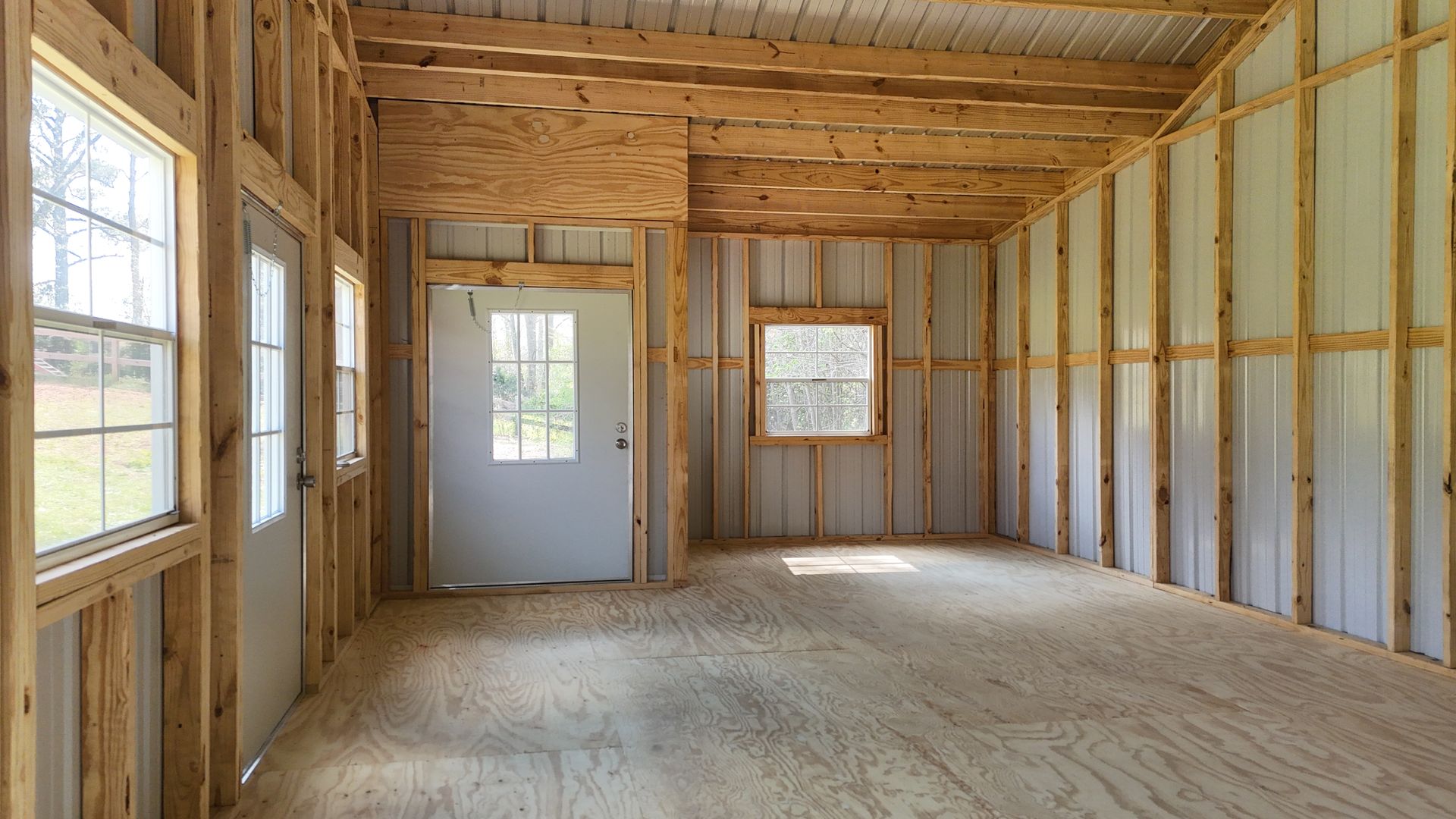 An unfinished interior room with exposed wooden studs, plywood flooring, metal wall panels, two doors, and several windows.