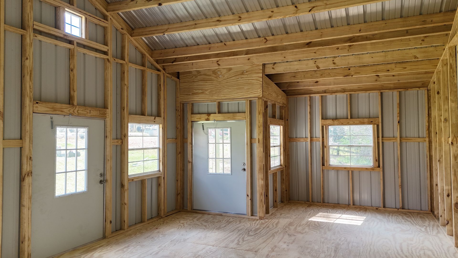 Interior view of a rustic wooden building under construction with exposed timber framing, two doors, and windows.