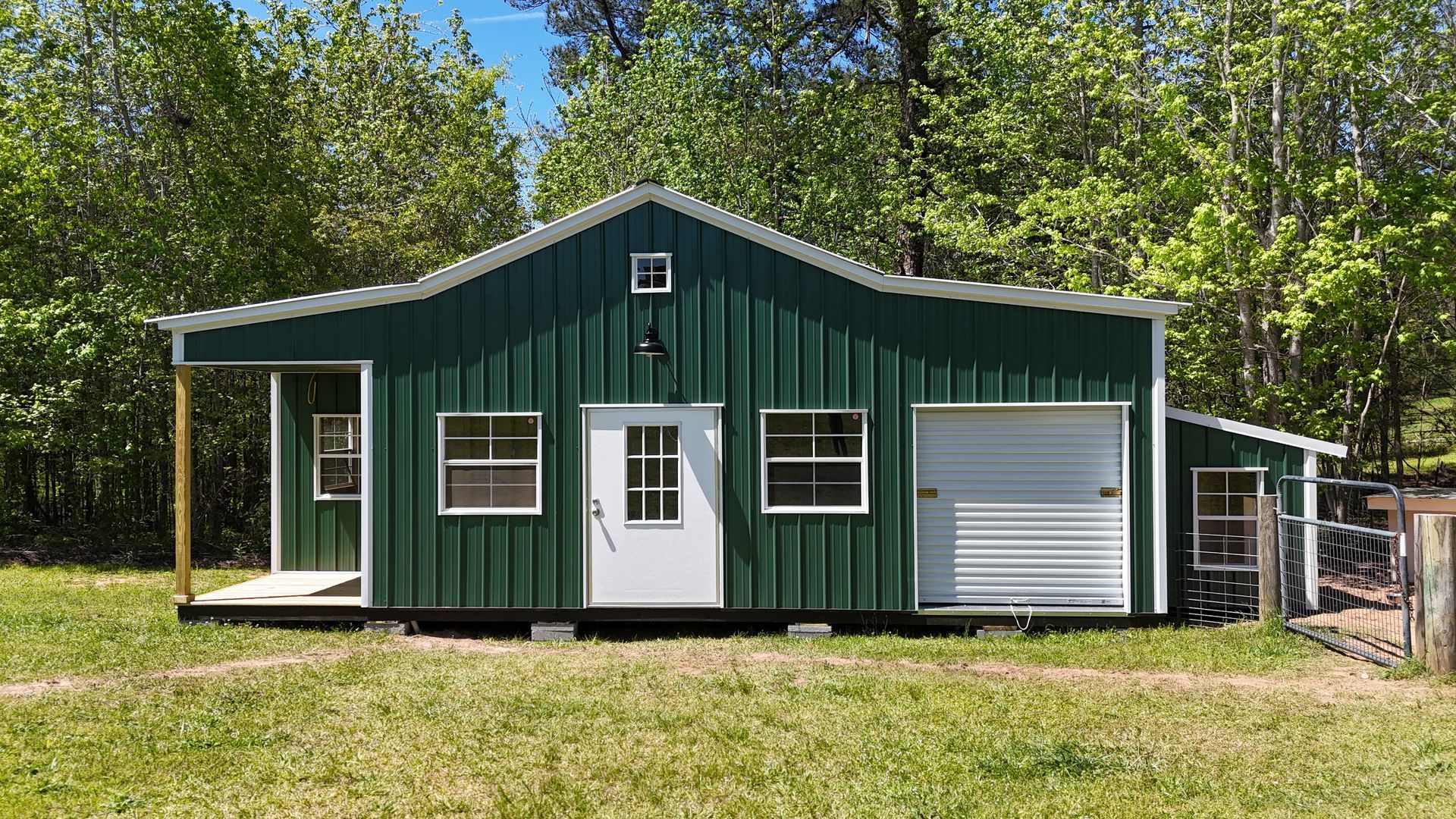 A forest-green metal workshop building with a white door, windows, and a side garage door, set in a grassy, wooded area.