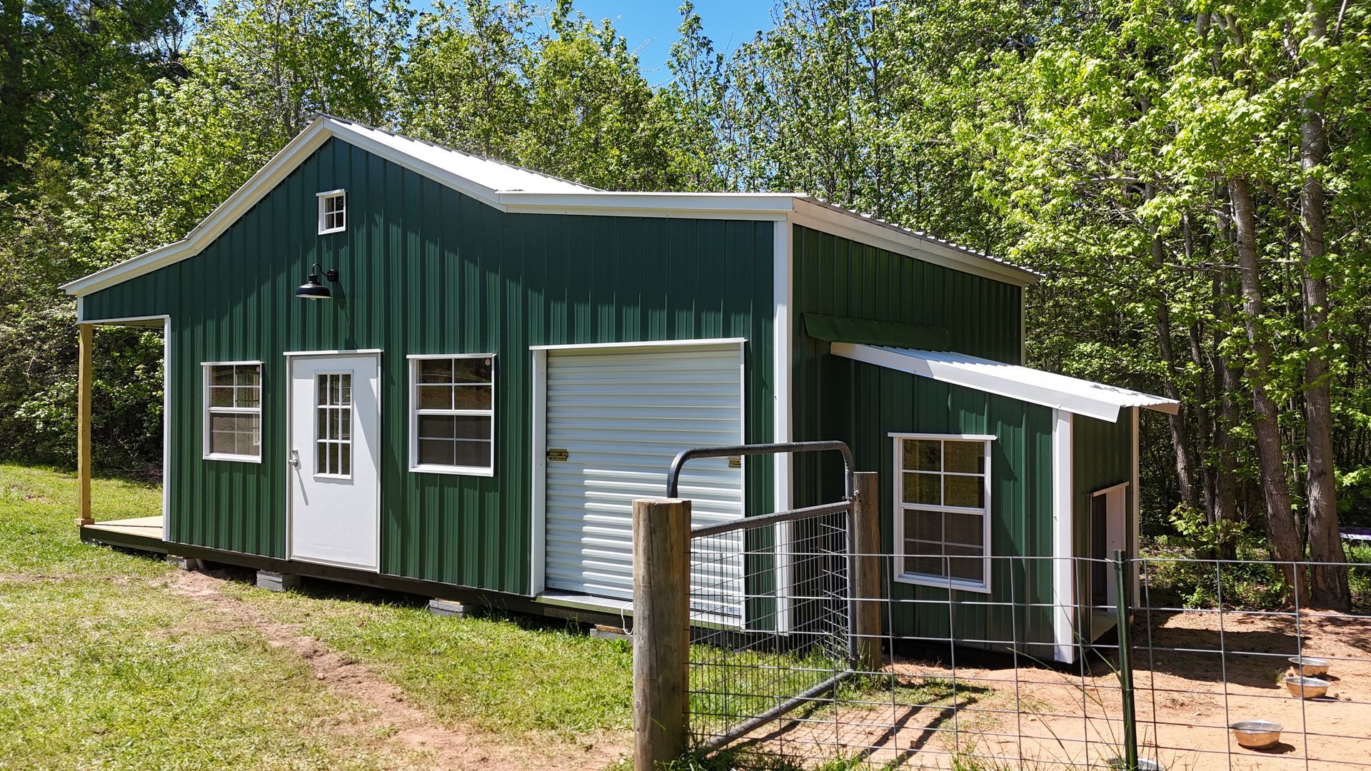 A green metal shed with a white door, garage door, windows, and a side overhang, situated in a wooded yard.