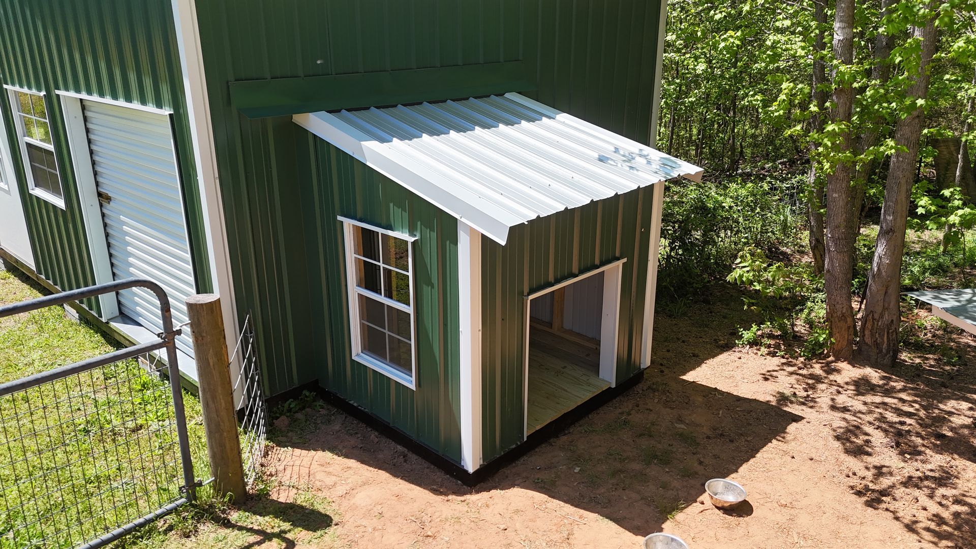 A green shed with a white-trimmed window and a silver-roofed alcove, situated outdoors near trees and a fenced area.