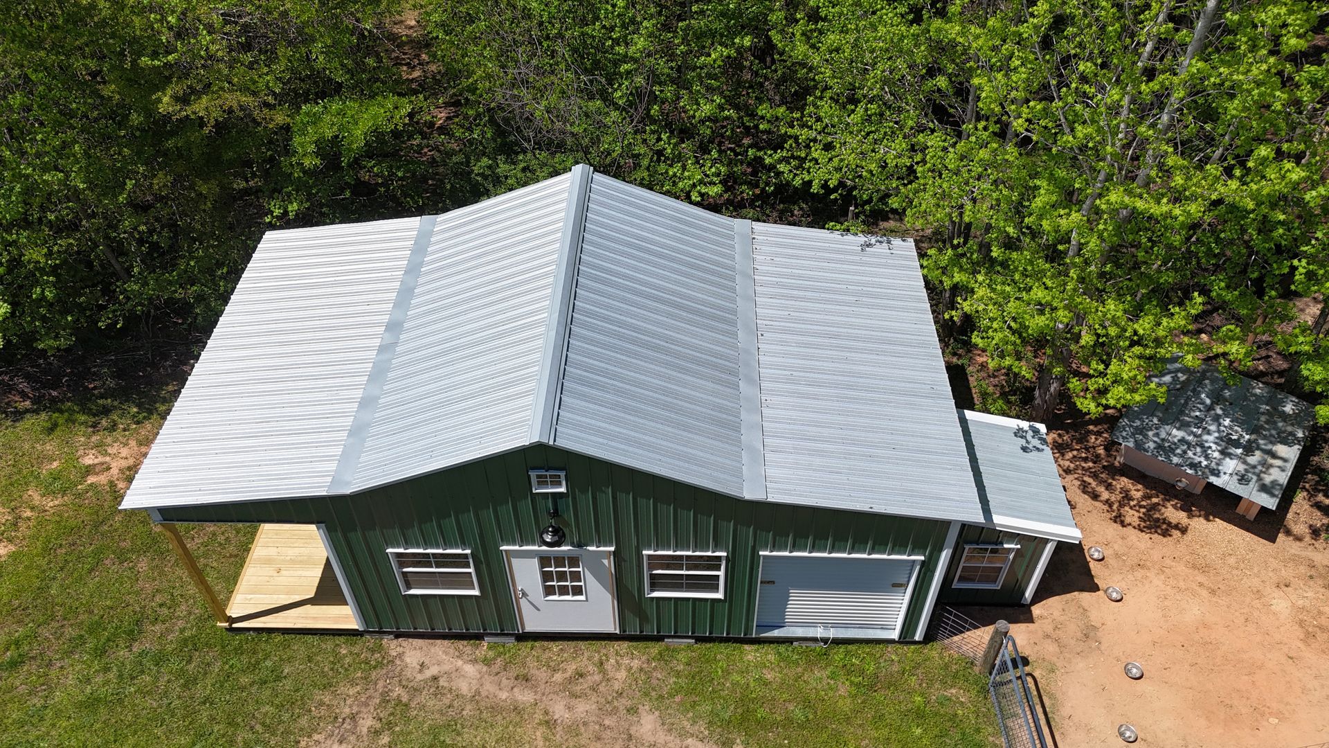 Aerial view of a small green shed with a metal roof, front porch, and garage door, surrounded by trees and grass.
