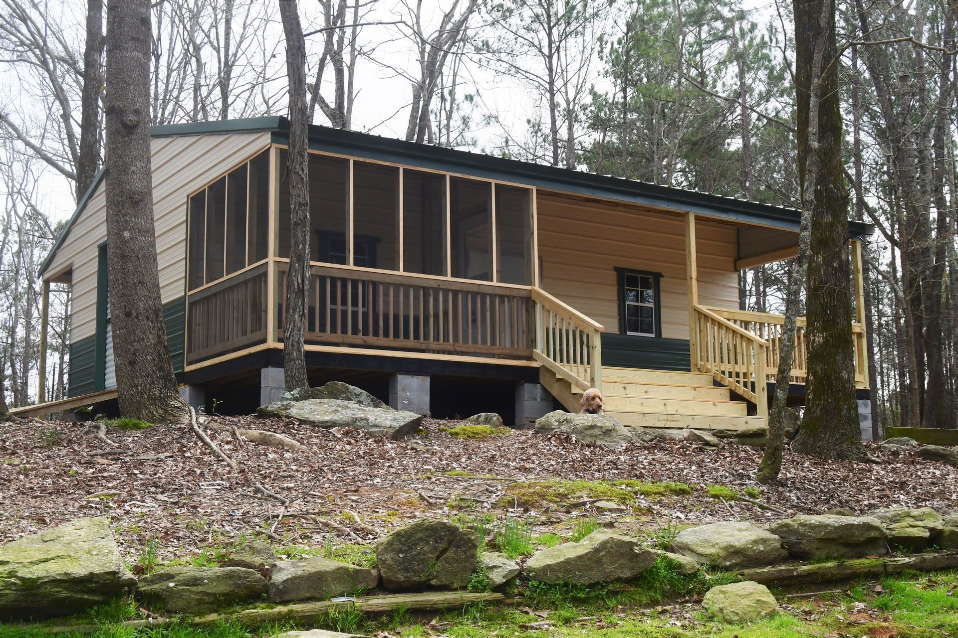 Cabin with screened porch and wooden deck in a wooded setting, on concrete blocks.