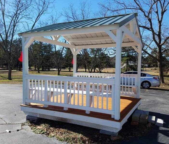 White gazebo with a green metal roof and wooden deck, set on a concrete base. Park setting with a car visible.