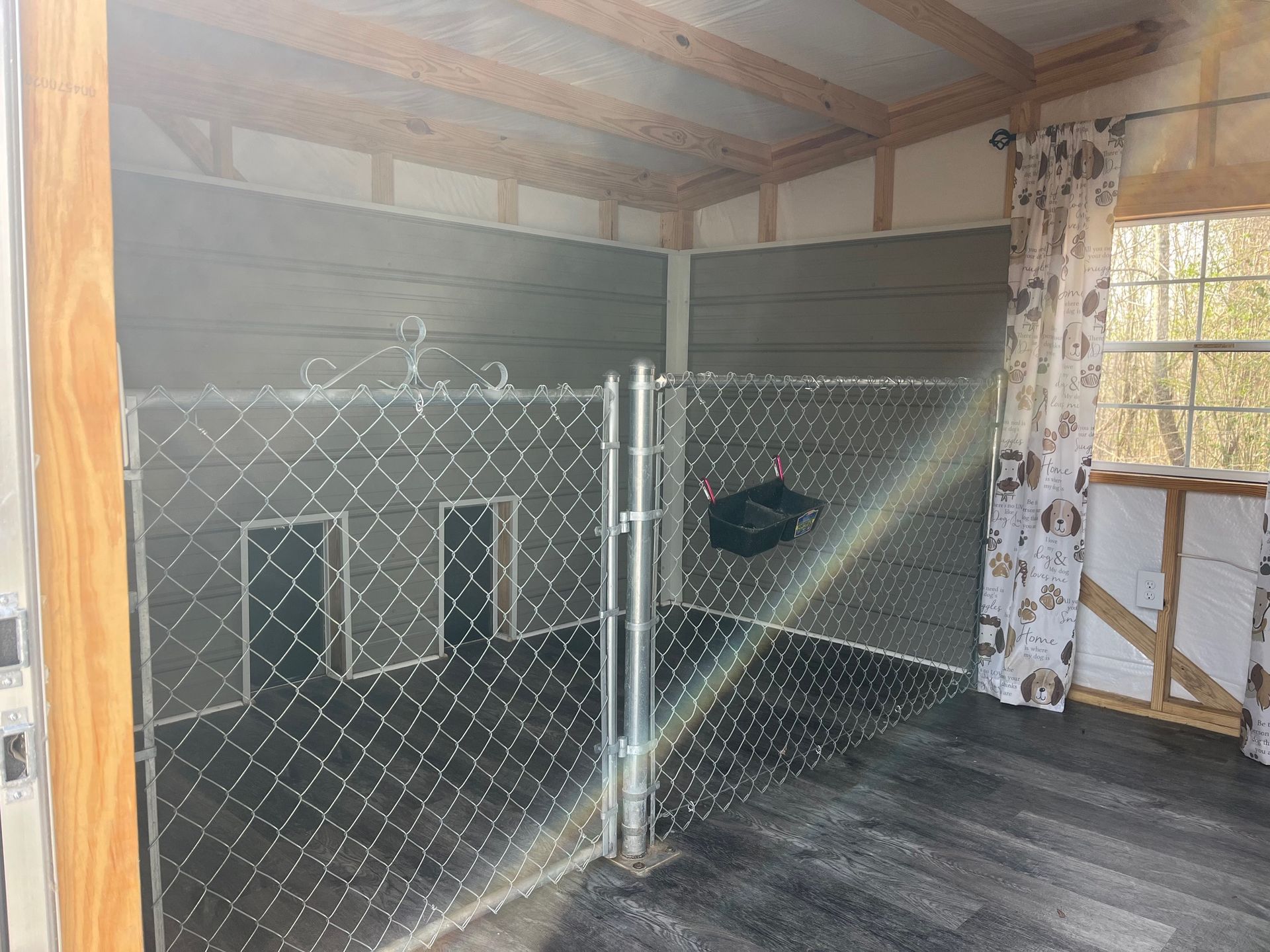Indoor dog kennel with chain-link fence, two dog doors, and a window with a dog-themed curtain.