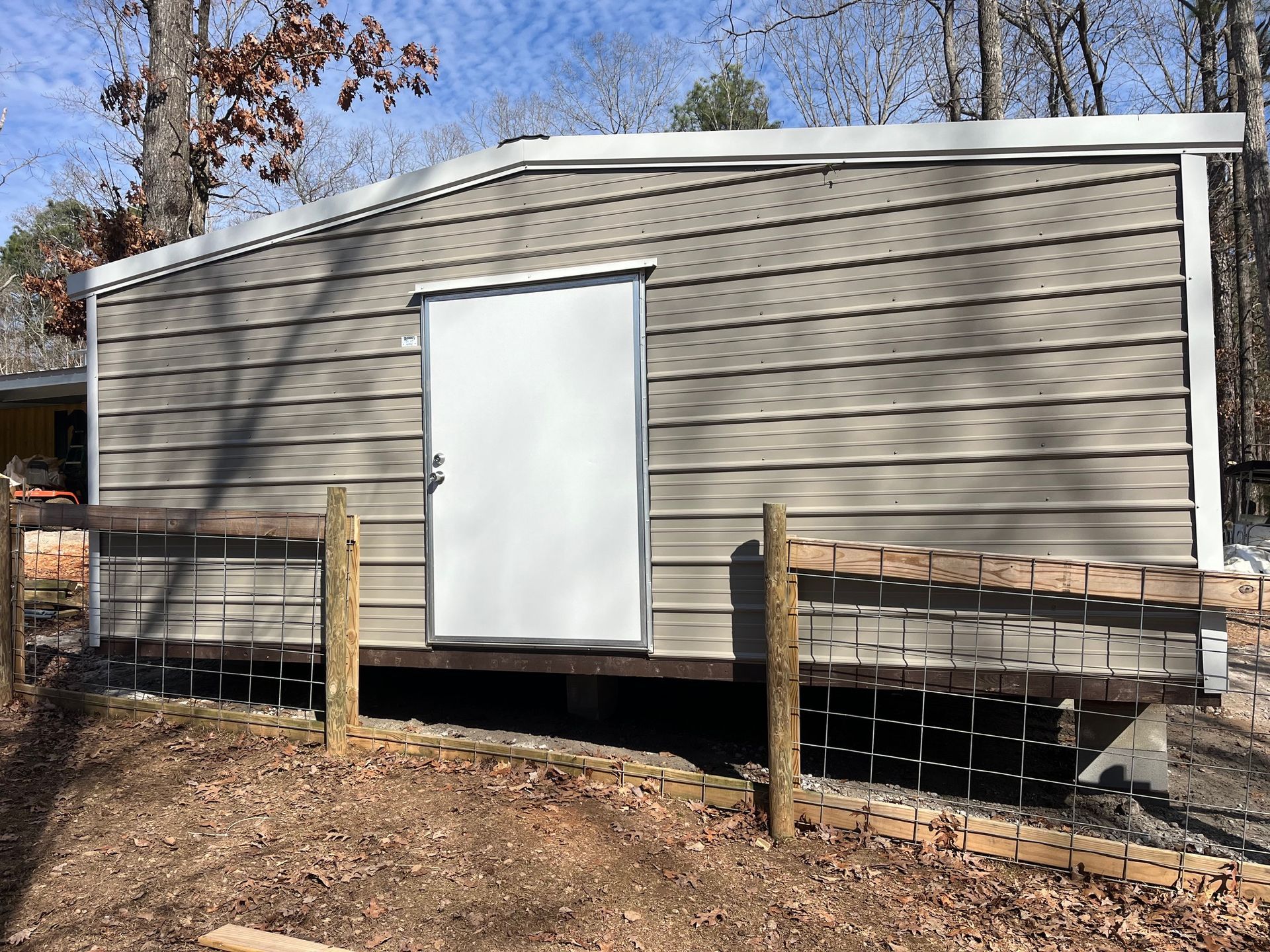 A small, metal-sided shed with a white door and wooden railing, set on a slightly elevated wooden platform.