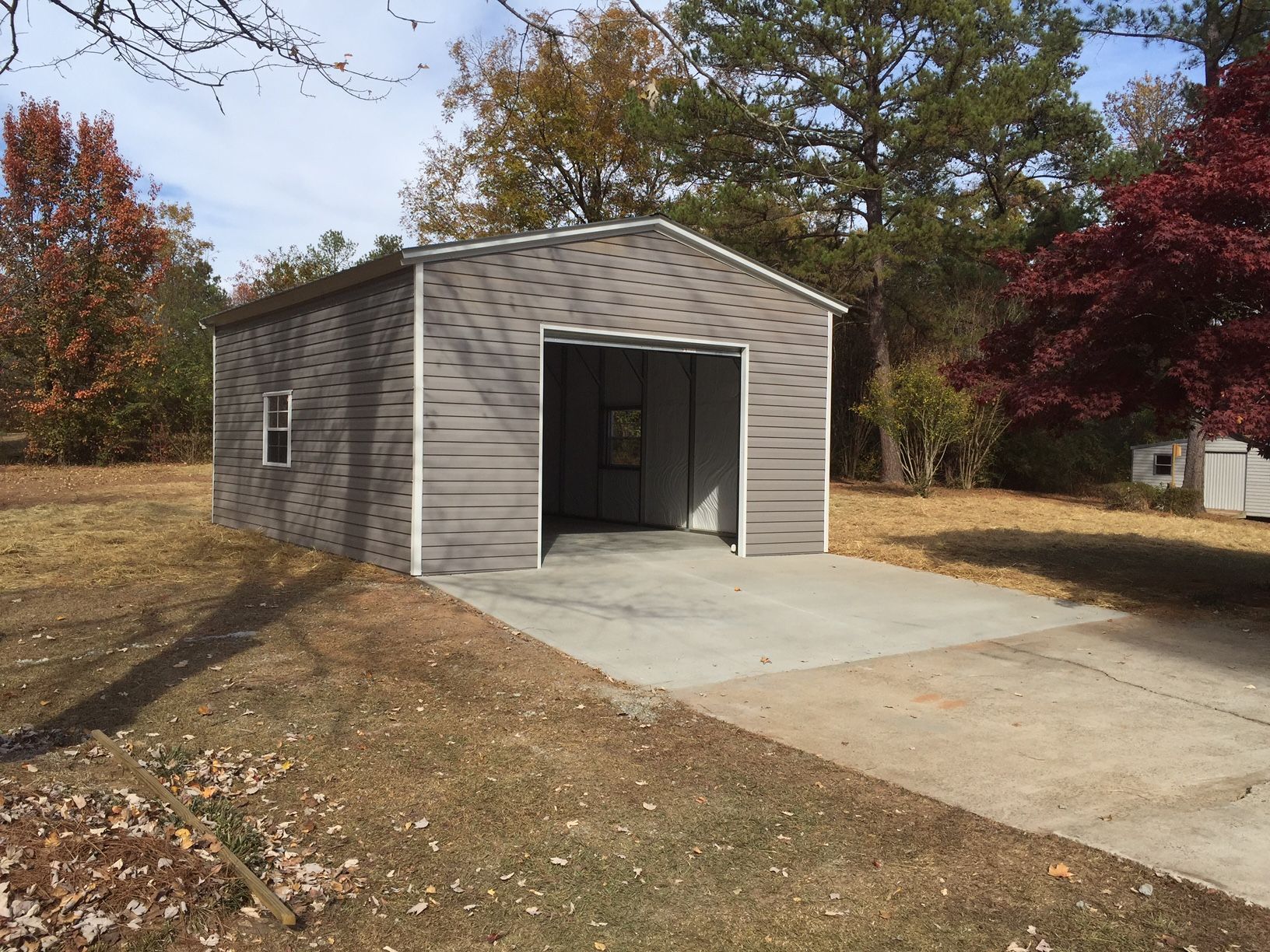 Gray metal garage with open door on a concrete pad, surrounded by dry grass and fall foliage.