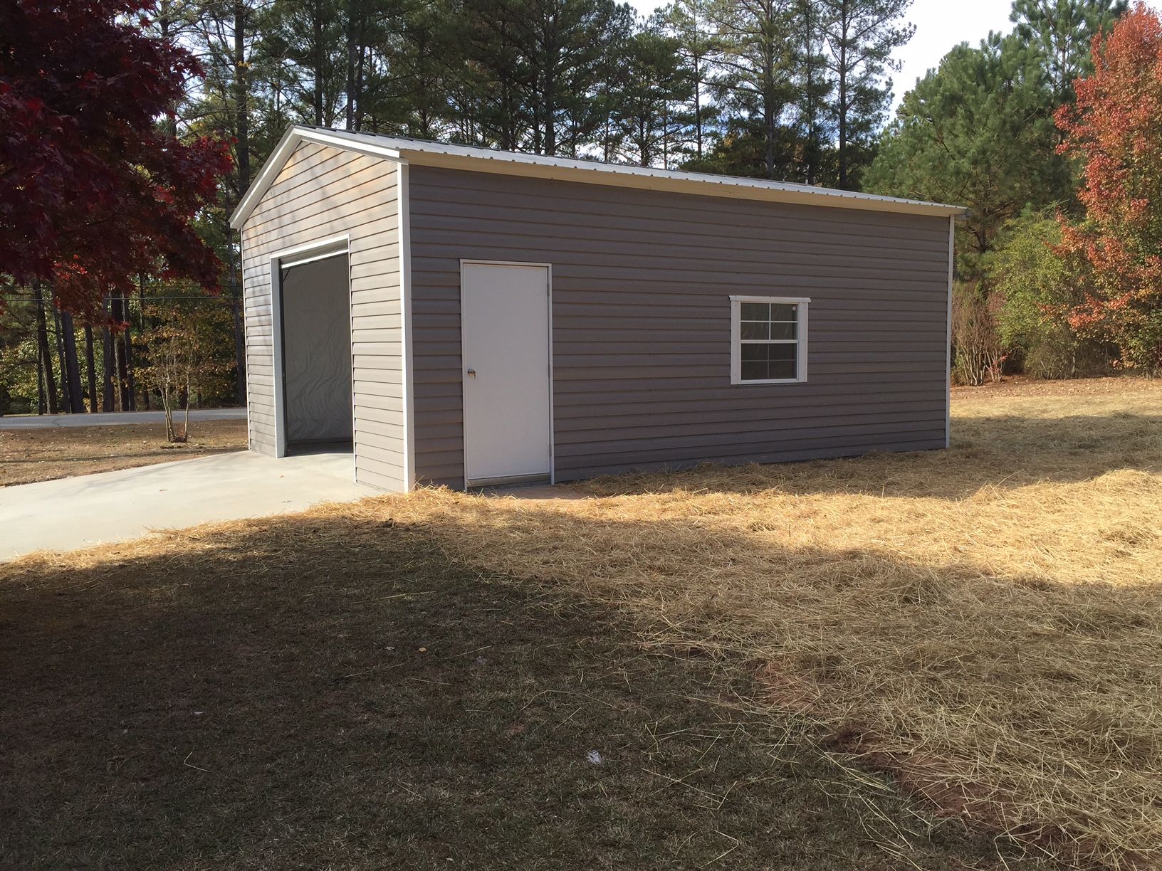 Metal gray storage building with an open garage door, white door, and window. Set on dry grass with a concrete driveway.