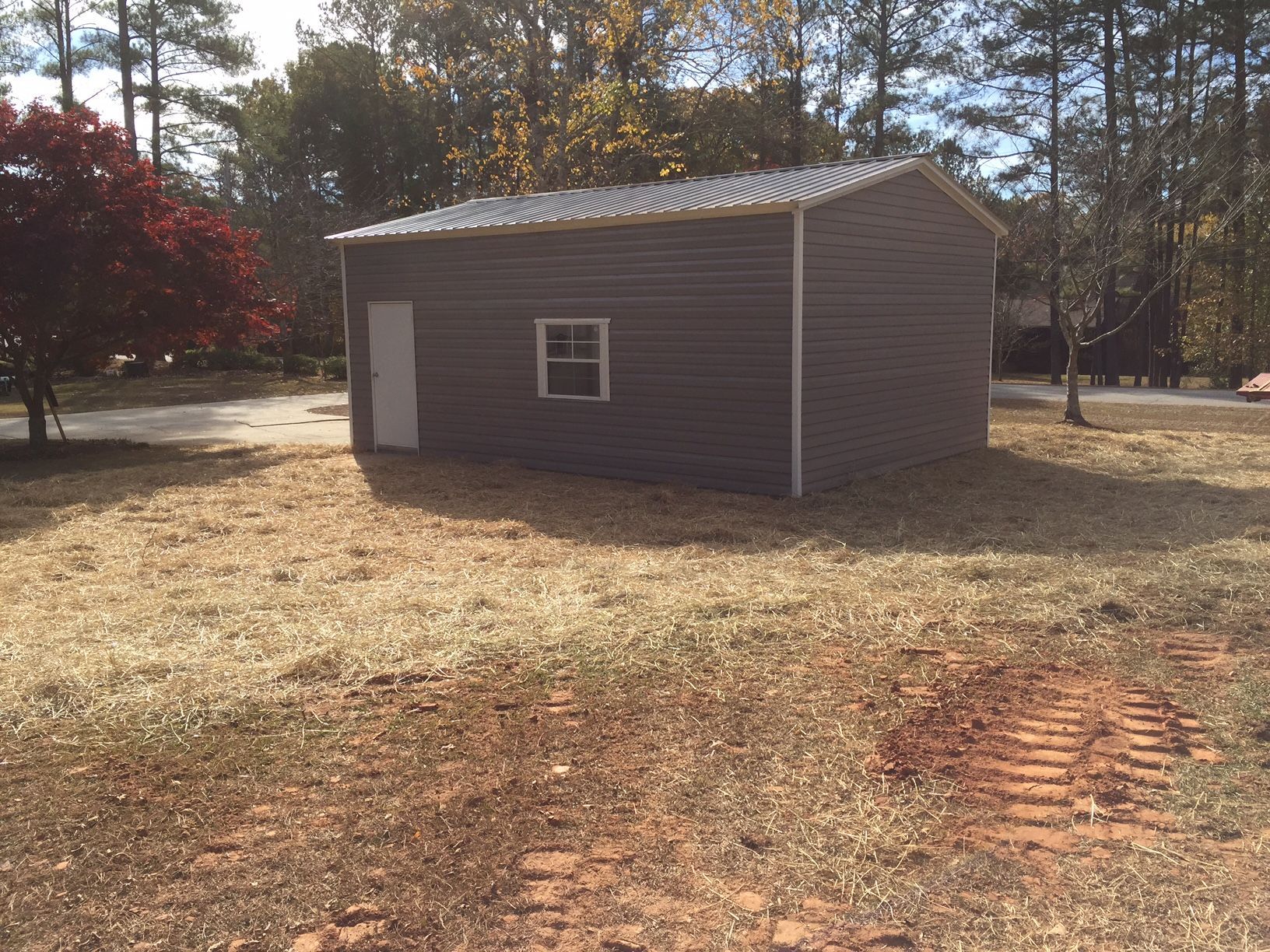 Gray metal shed with a white door and window, set in a yard with sparse grass.