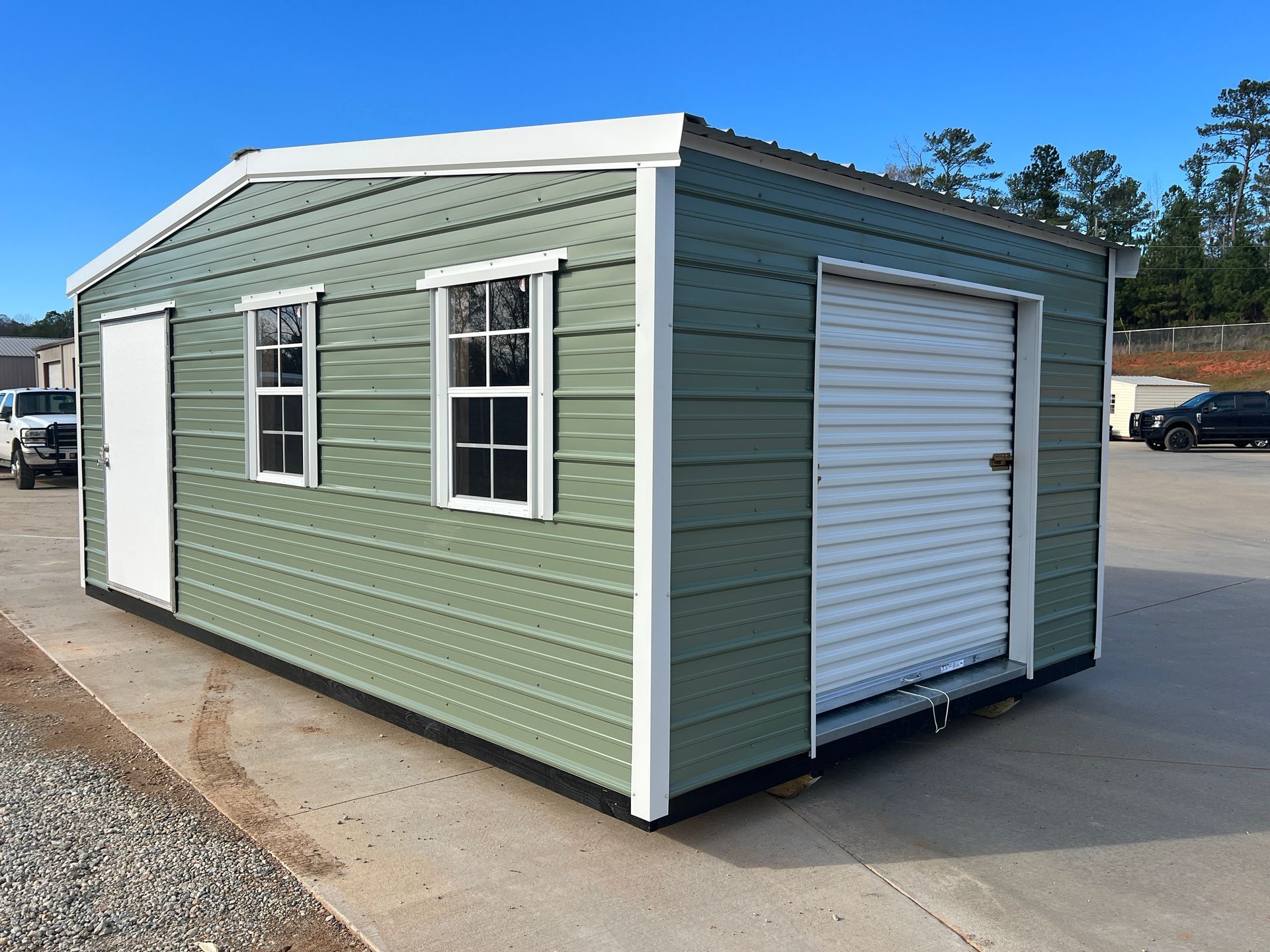 Green metal storage building with white door, window frames, and garage door on a concrete surface.