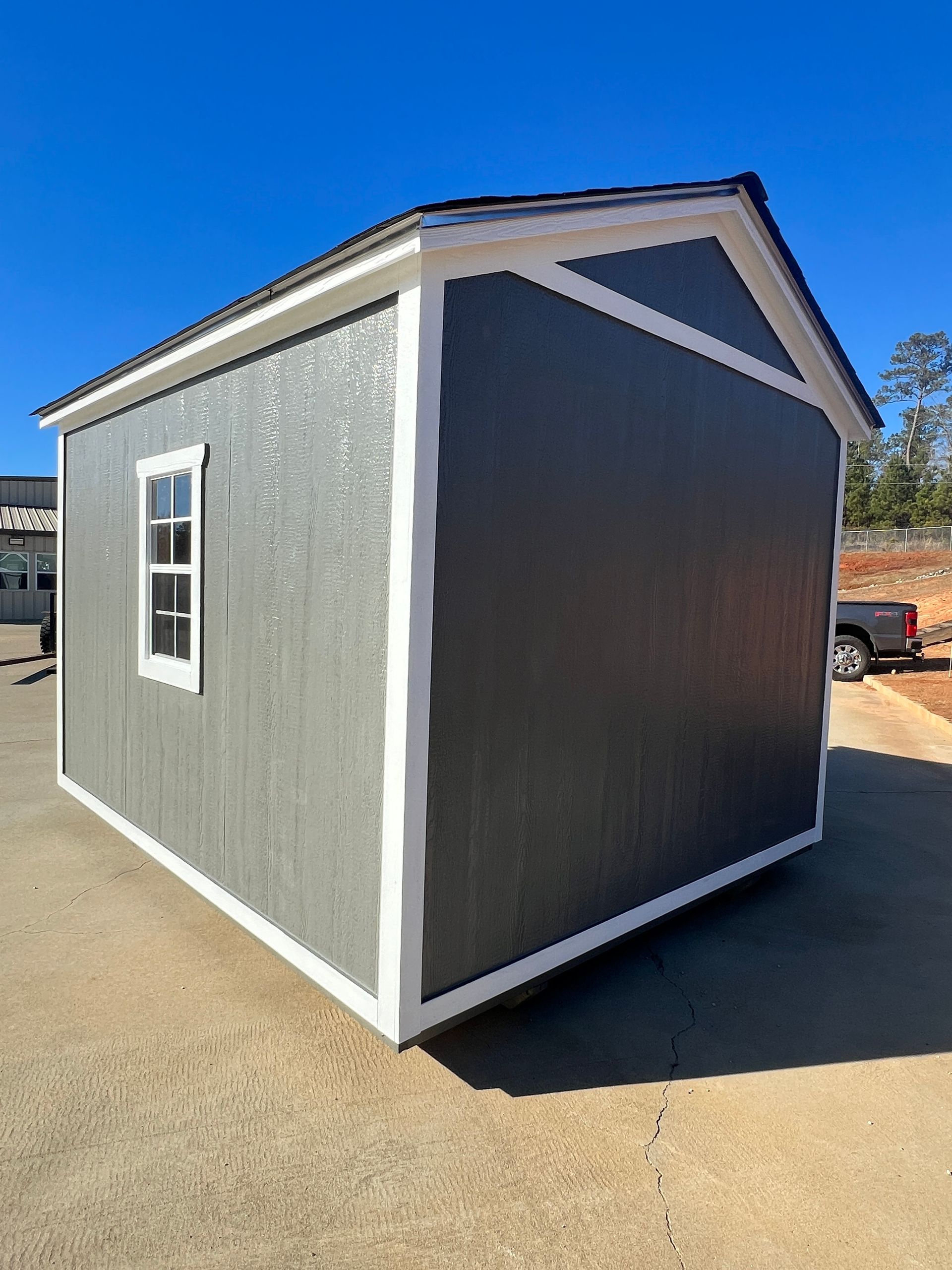 Gray shed with white trim, window, and black roof, on concrete in daylight.