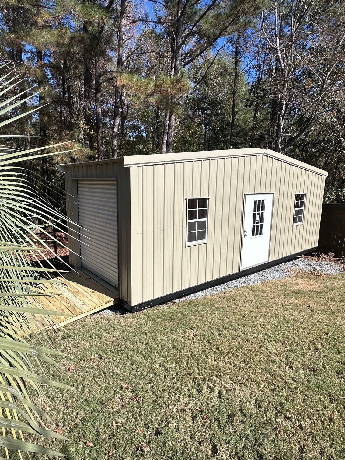 Beige shed with a roll-up door, windows, and white door, set in a yard with a grassy area.