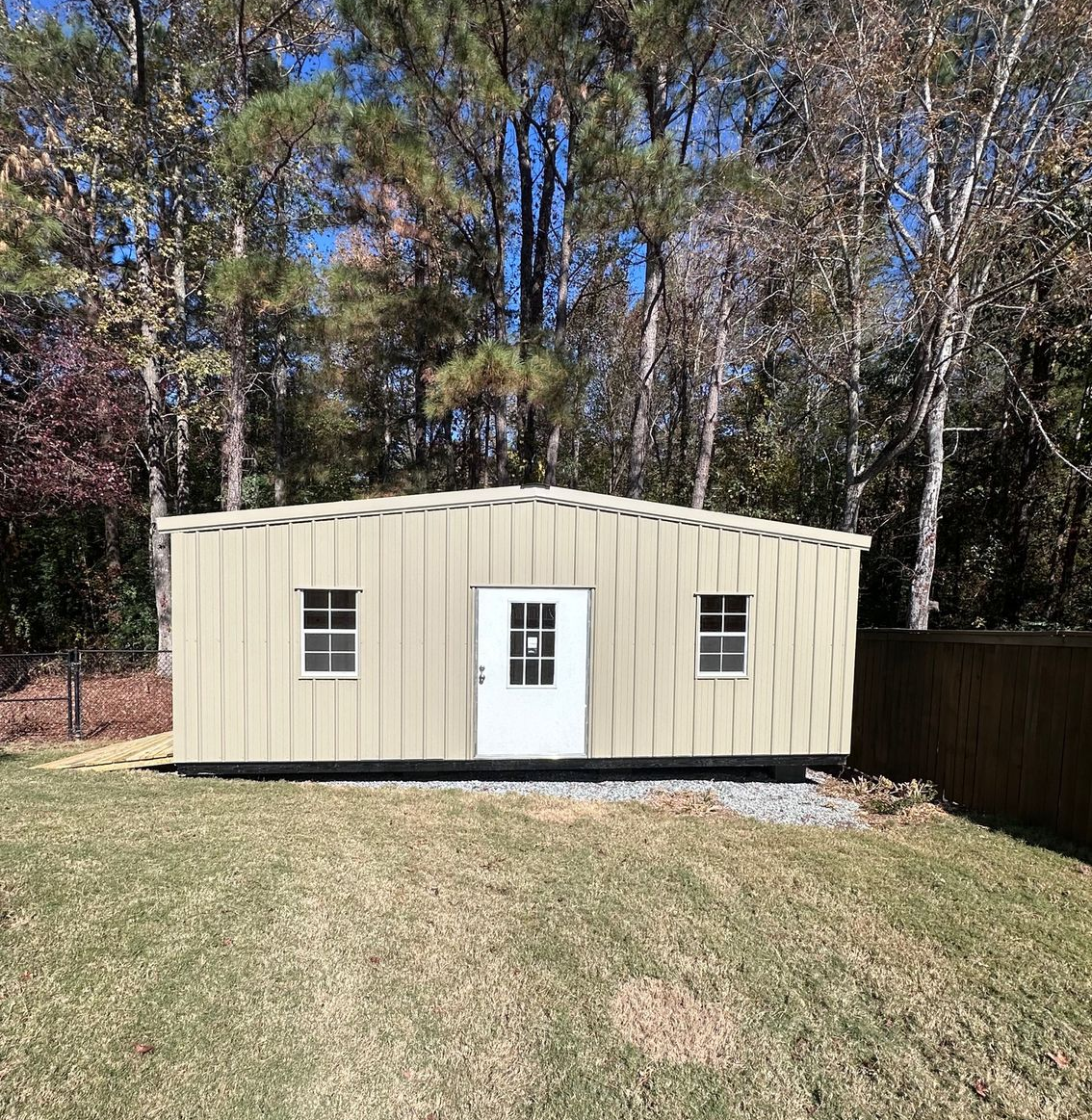 Tan shed with white door and two windows, set in a backyard with trees and grass.