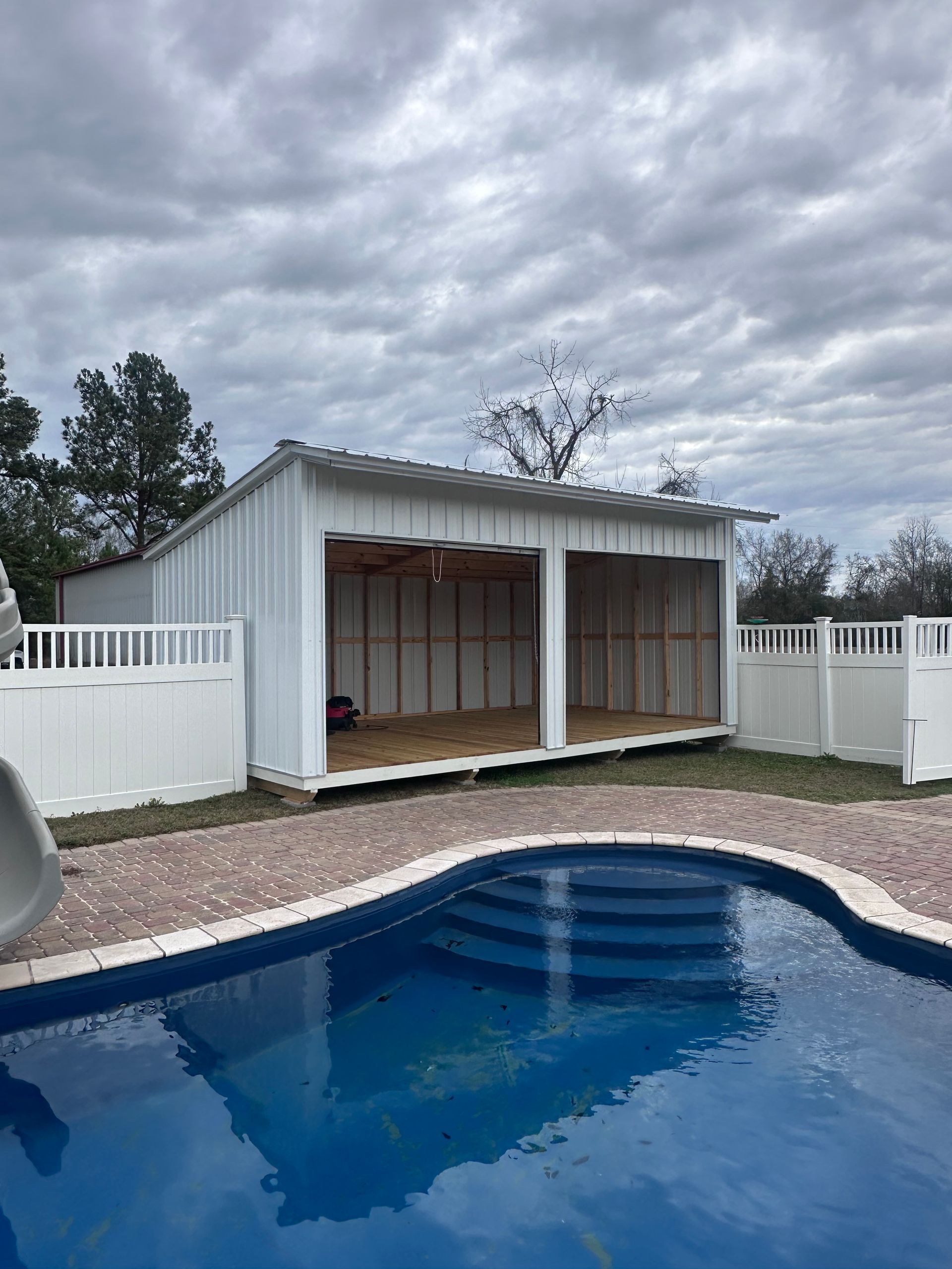 Poolside shed with open bays, white walls, and a corrugated metal roof. A cloudy sky is in the background.