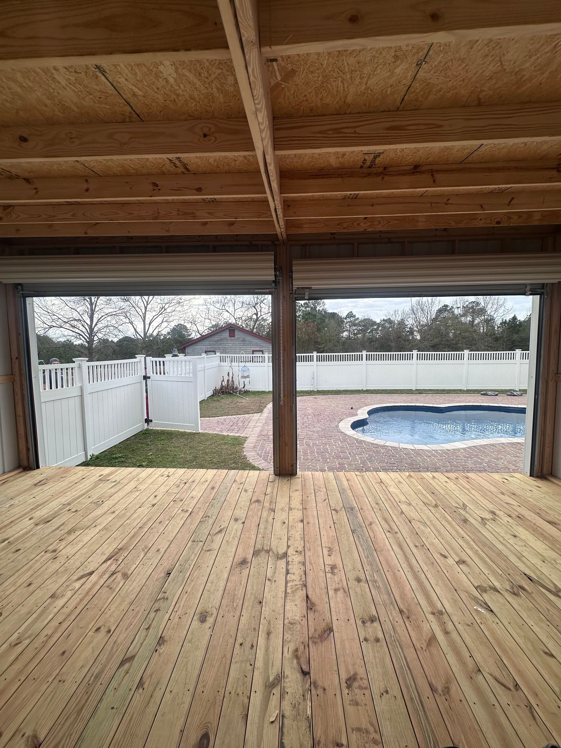 View from a wooden deck, through openings, to a backyard with a pool and white fence.