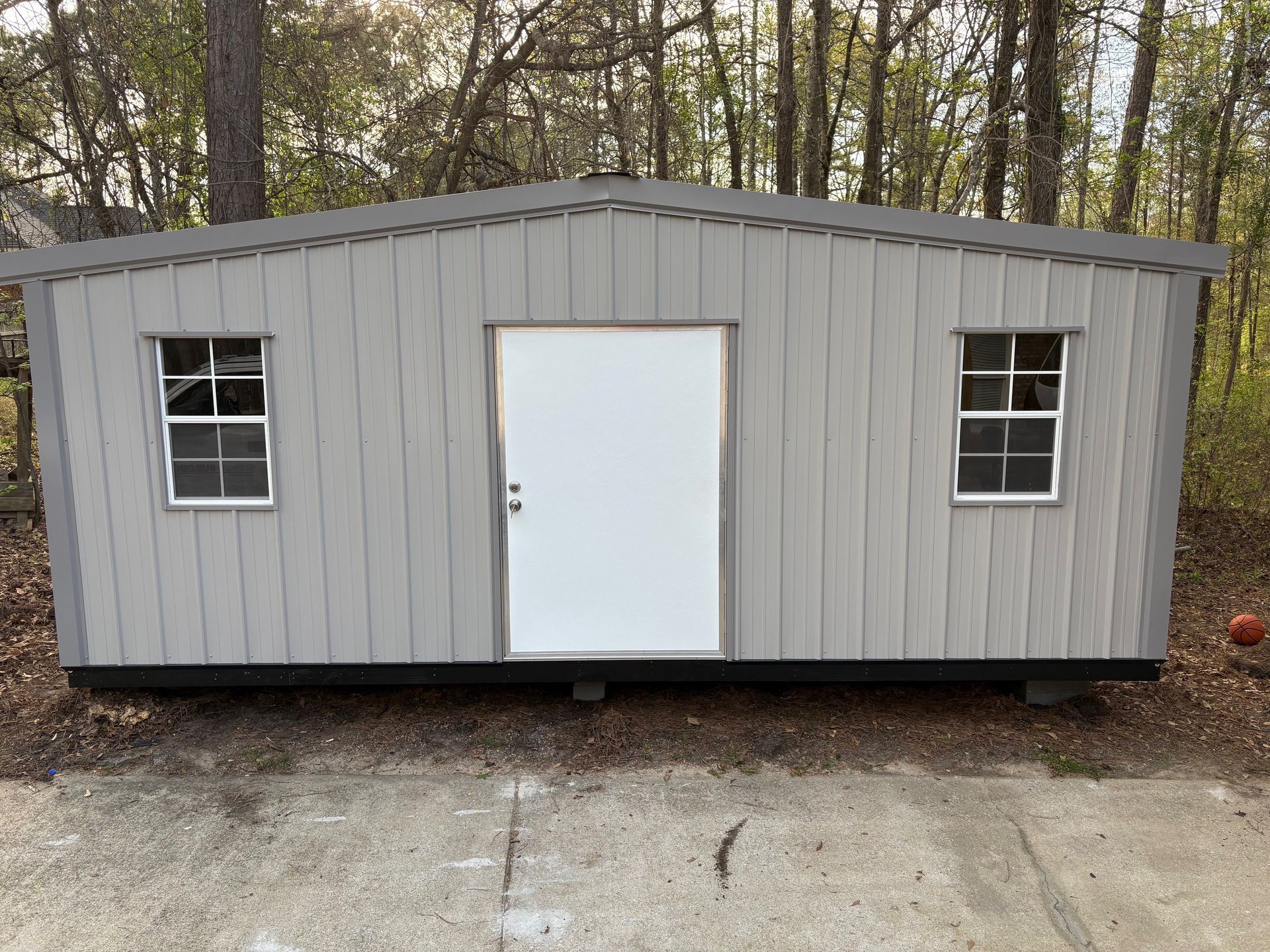 A grey, rectangular storage shed with a centered white door and two white-framed windows, positioned on a concrete pad.