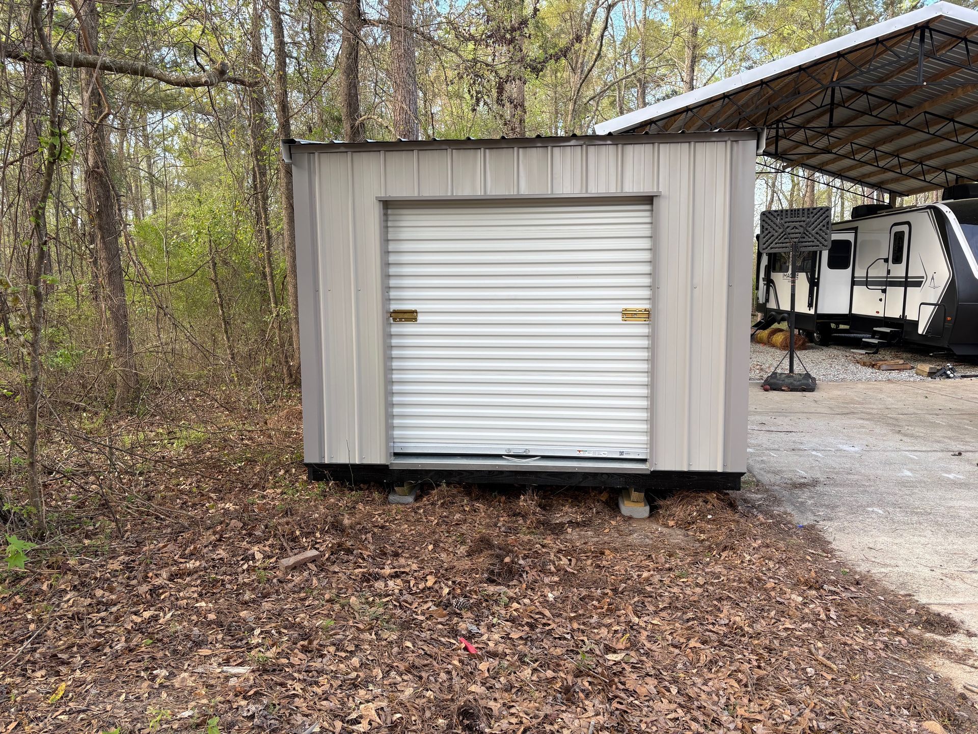 A light gray metal storage shed with a roll-up door sits in a wooded area next to a gravel driveway and a parked RV.