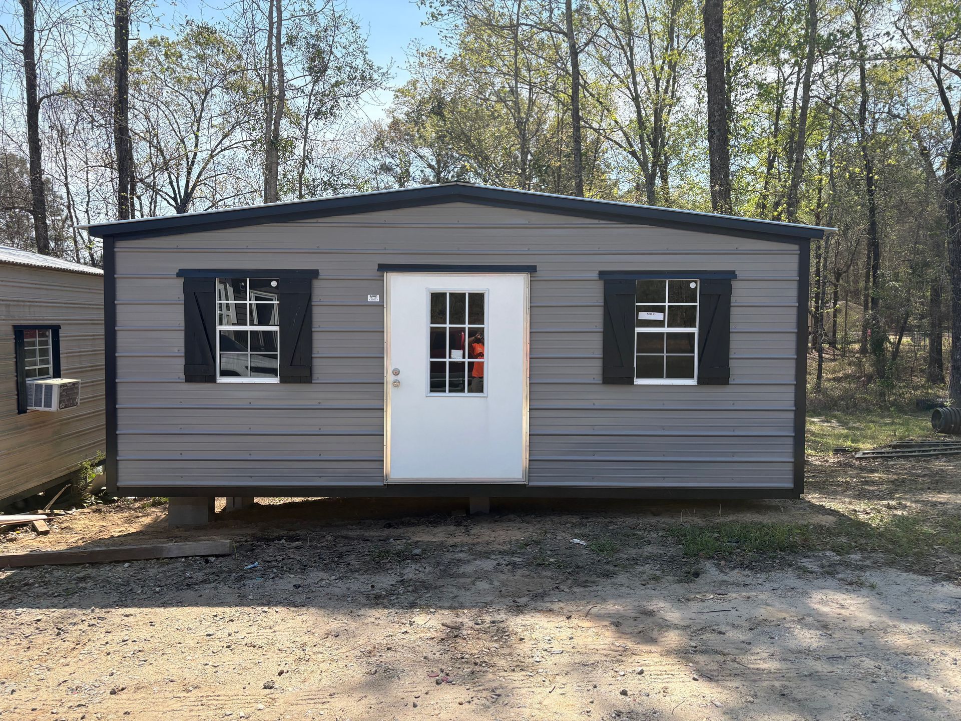 A grey portable shed with a white door, two windows with black shutters, and black trim, set in a wooded area.