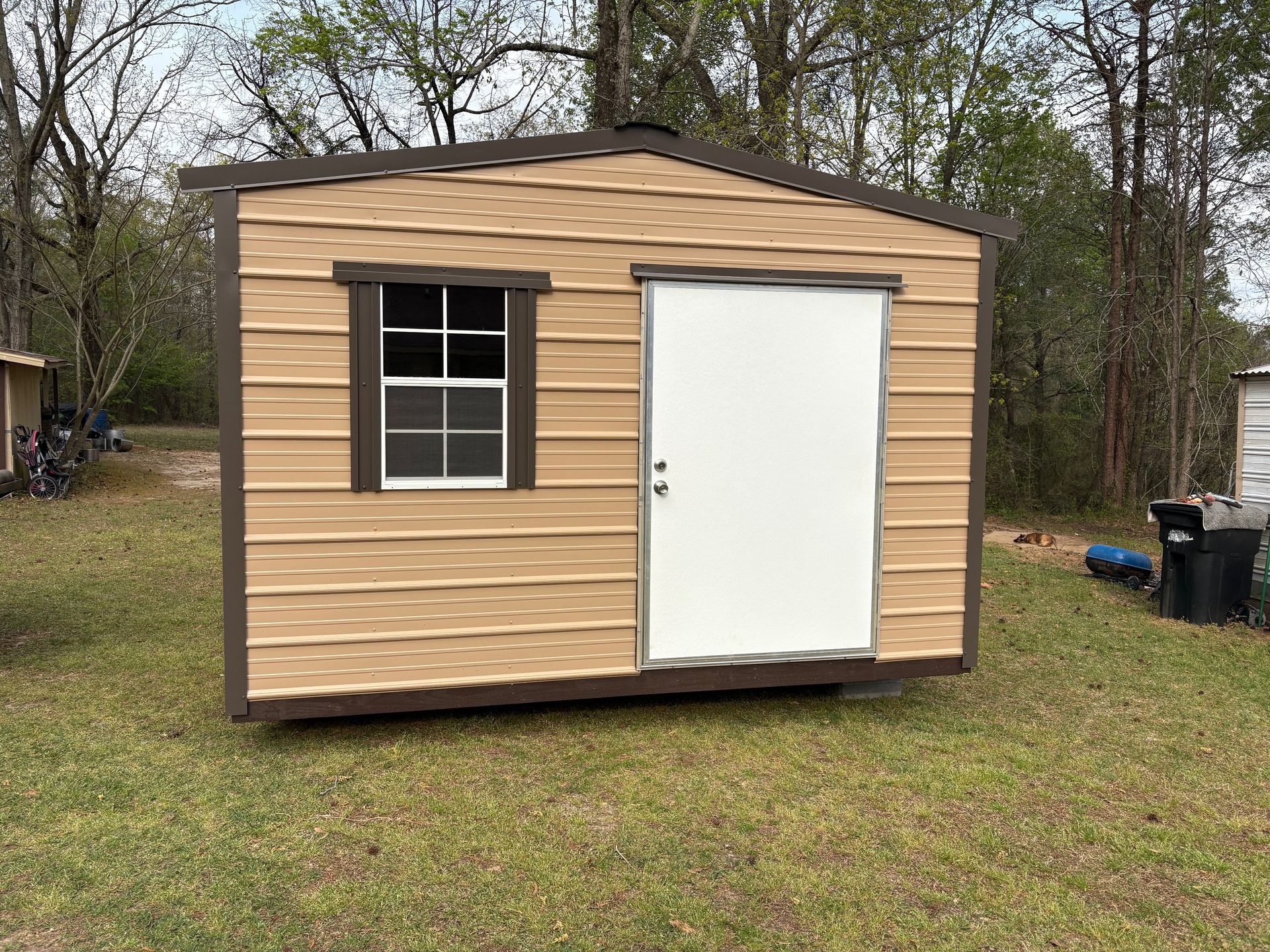 A tan-sided storage shed with dark brown trim, a window with a white grid, and a solid white door in a grassy area.
