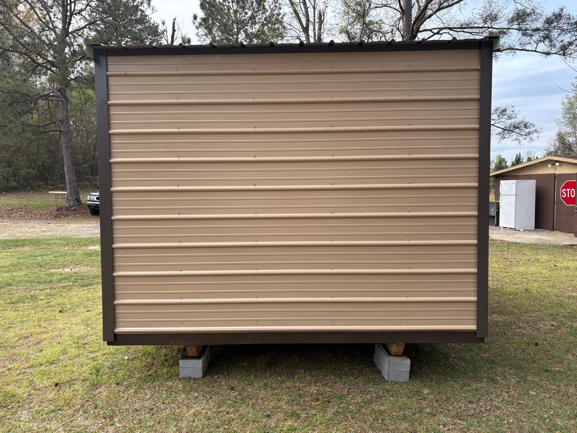 A beige metal shed exterior on cinder block foundation, situated in a grassy lot with trees in the background.