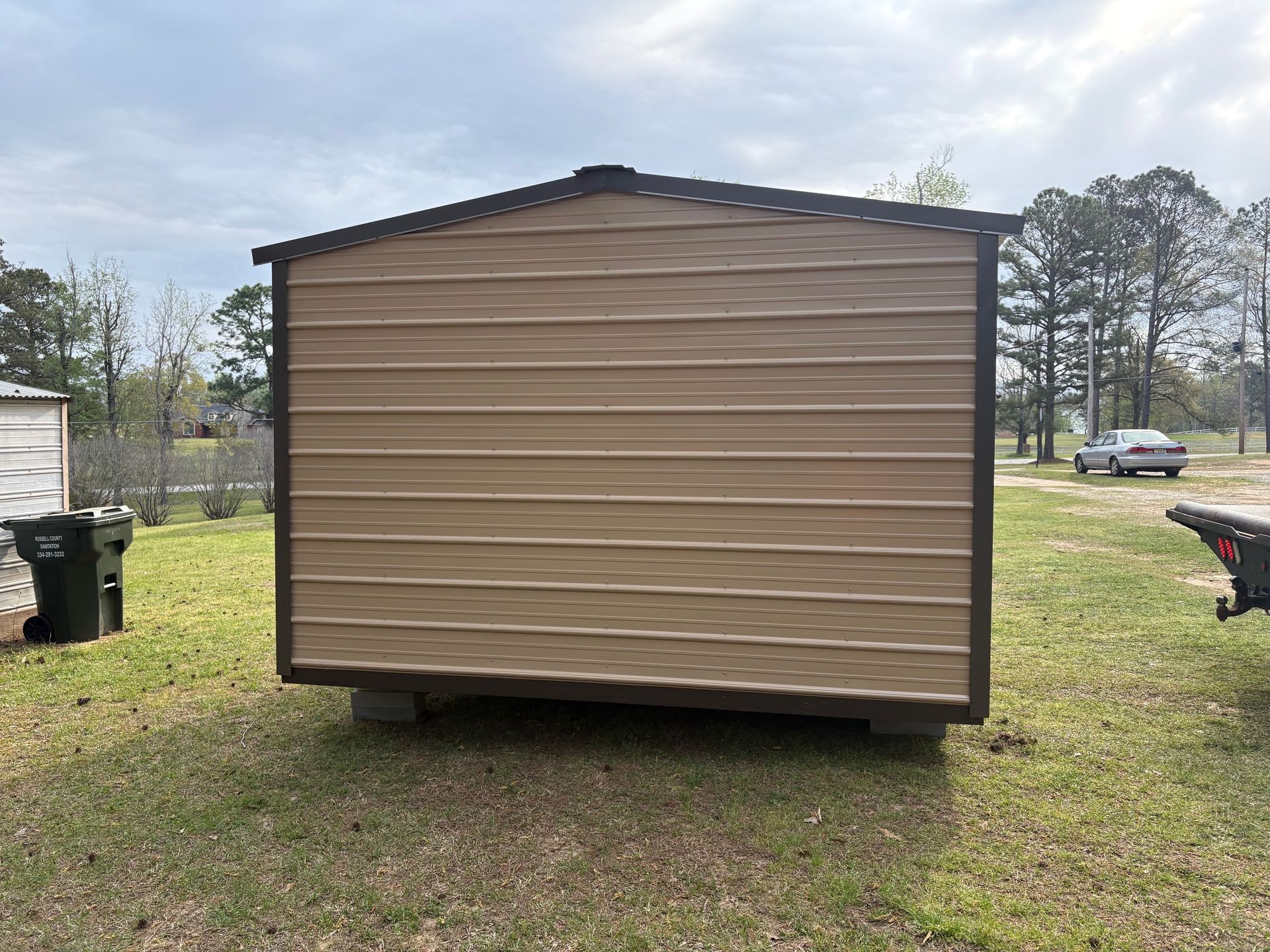 A rectangular storage shed with tan horizontal siding and dark brown trim stands on a grassy lot under a cloudy sky.