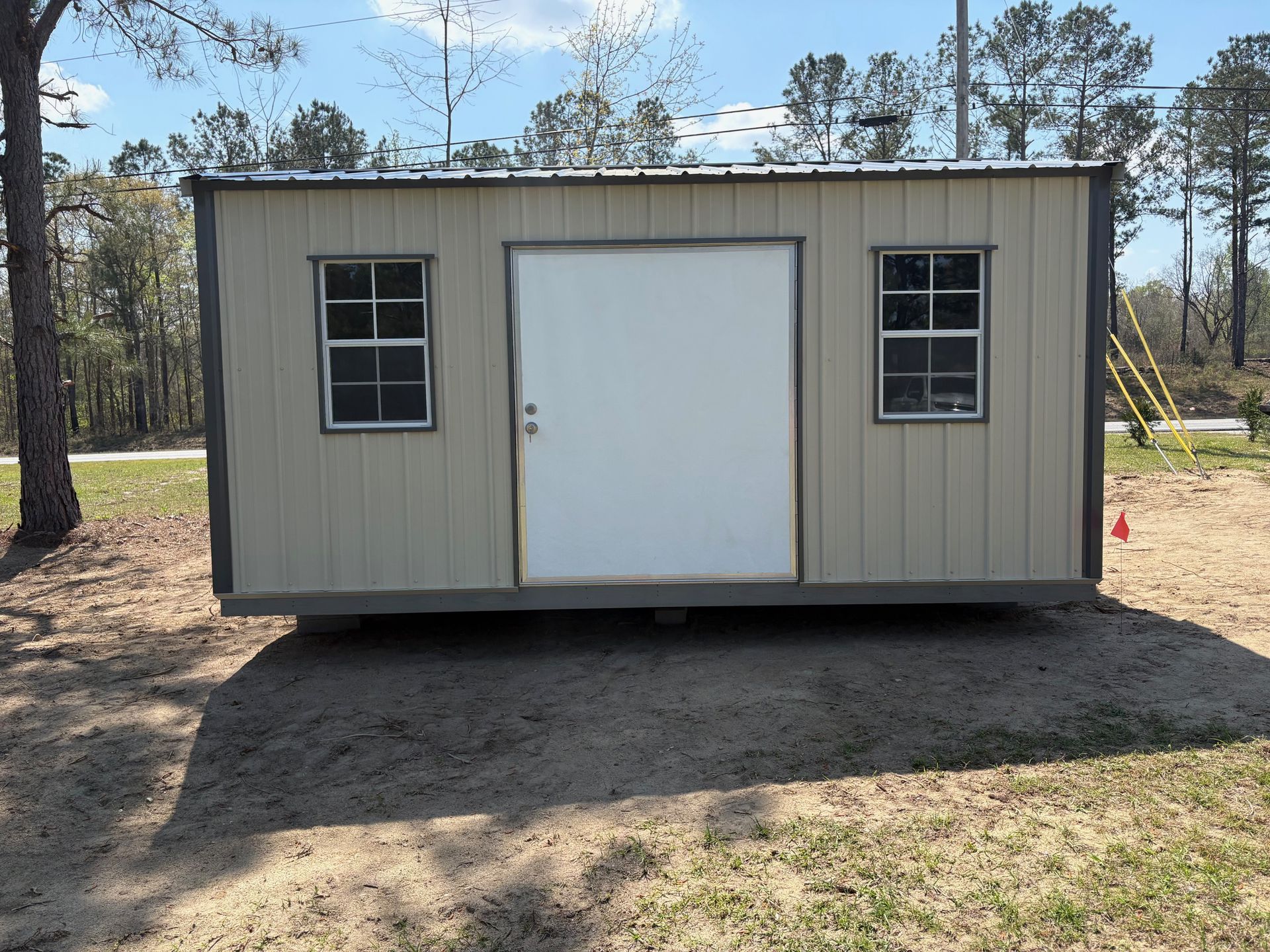 A tan, rectangular portable storage shed with a white central door and two matching grid windows on a sandy lot.