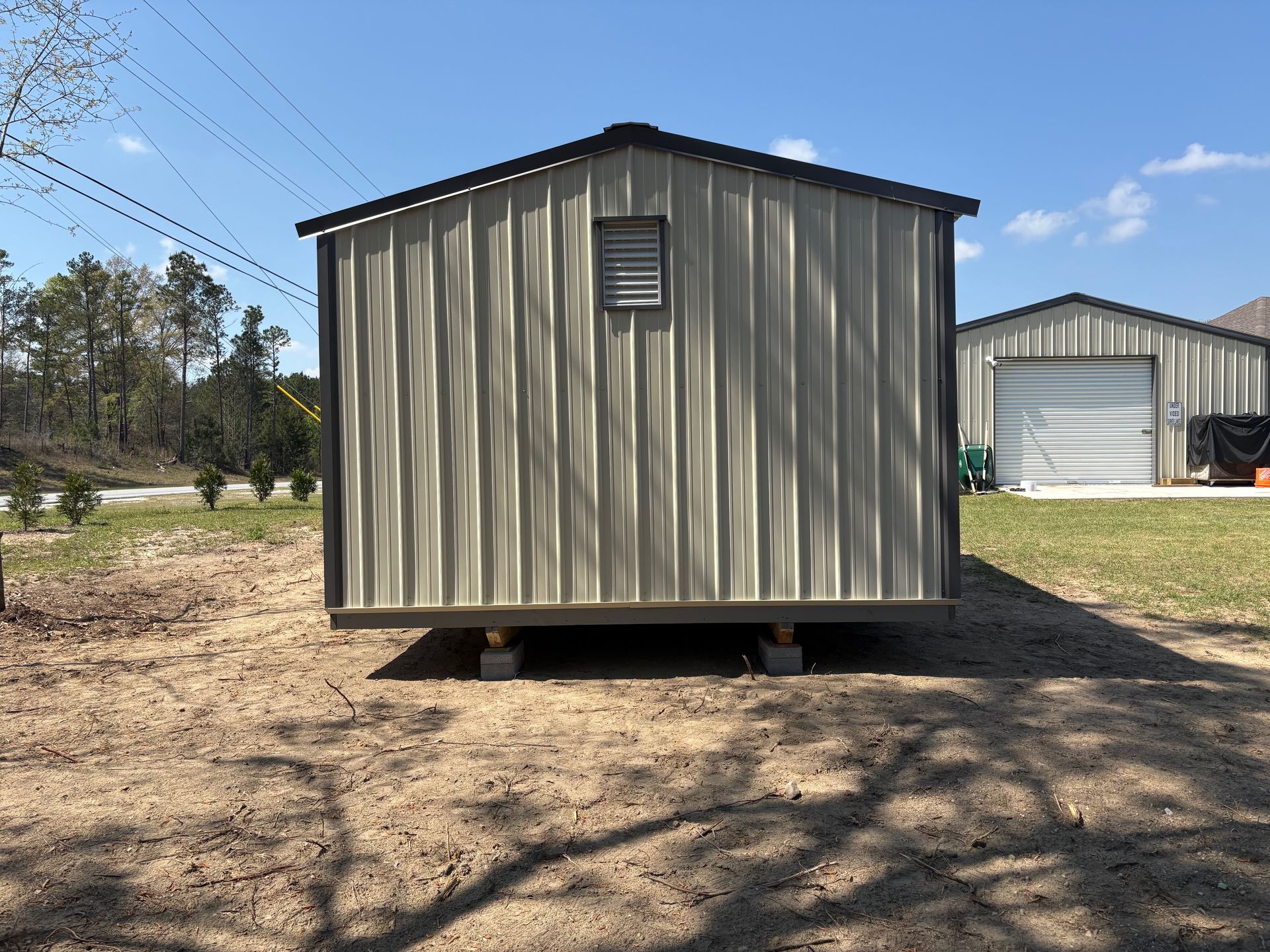 A tan metal shed sits on concrete blocks in a grassy yard, with a larger garage building in the background.