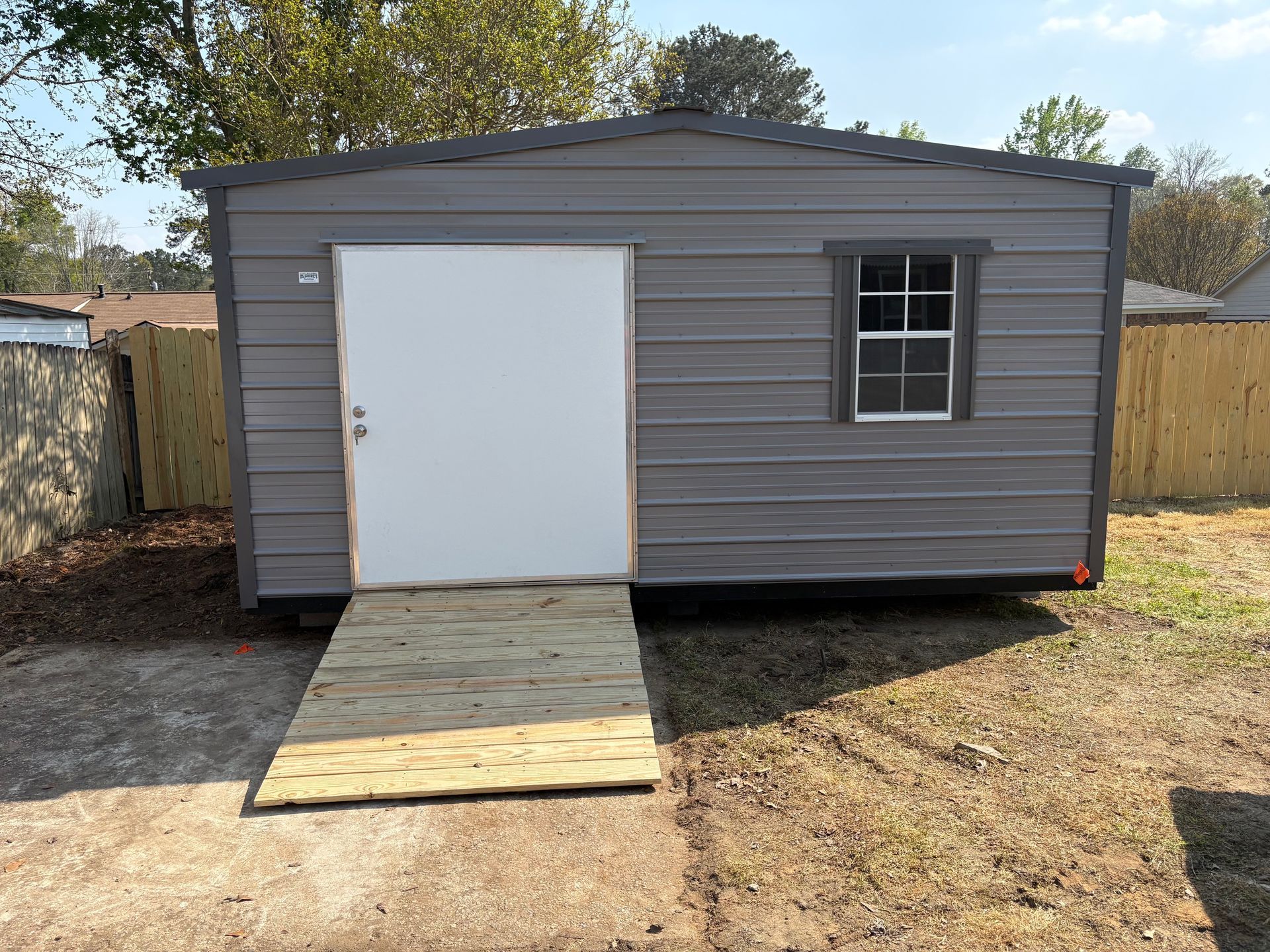 A grey storage shed with a white door and a single window, featuring a wooden ramp leading to the entrance.