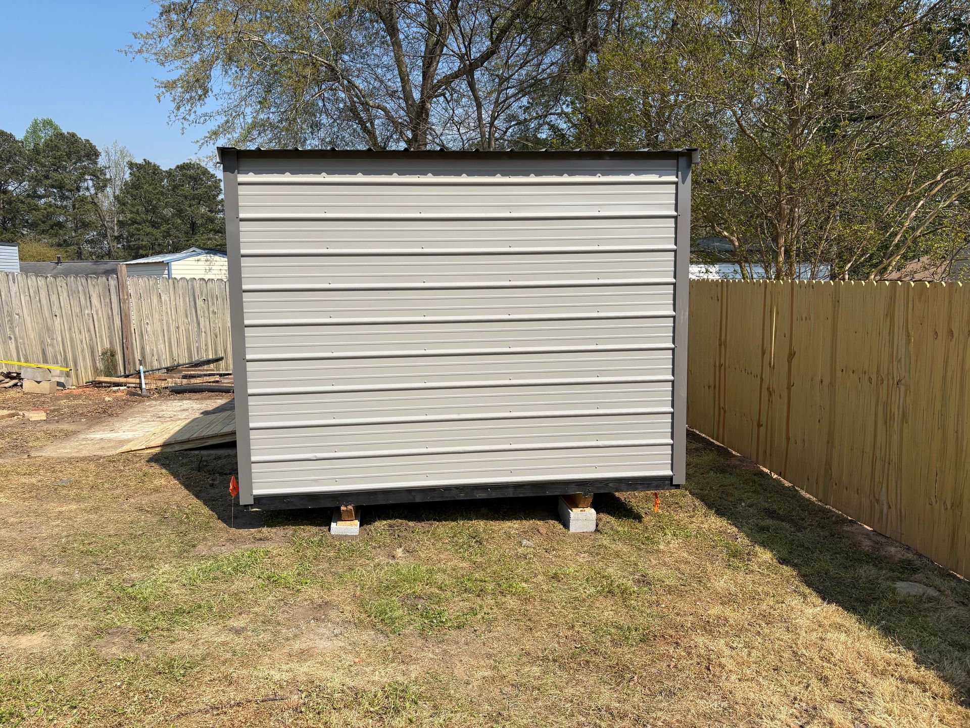 A light-gray metal storage shed stands on concrete blocks in a grassy yard between two fences.