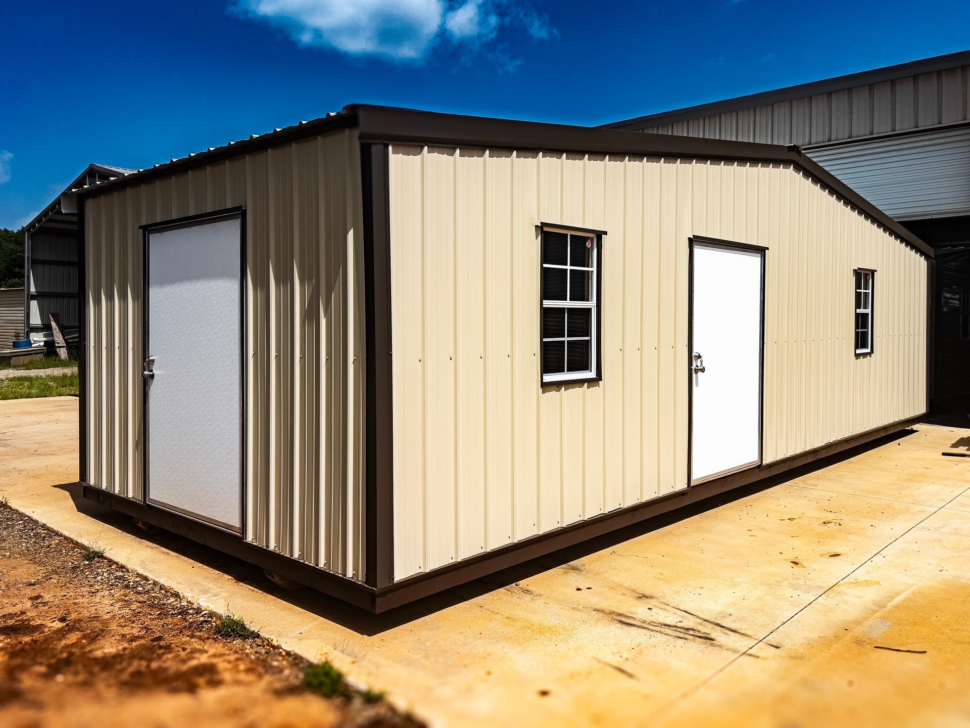 A metal building with two doors and two windows is sitting on top of a dirt driveway.