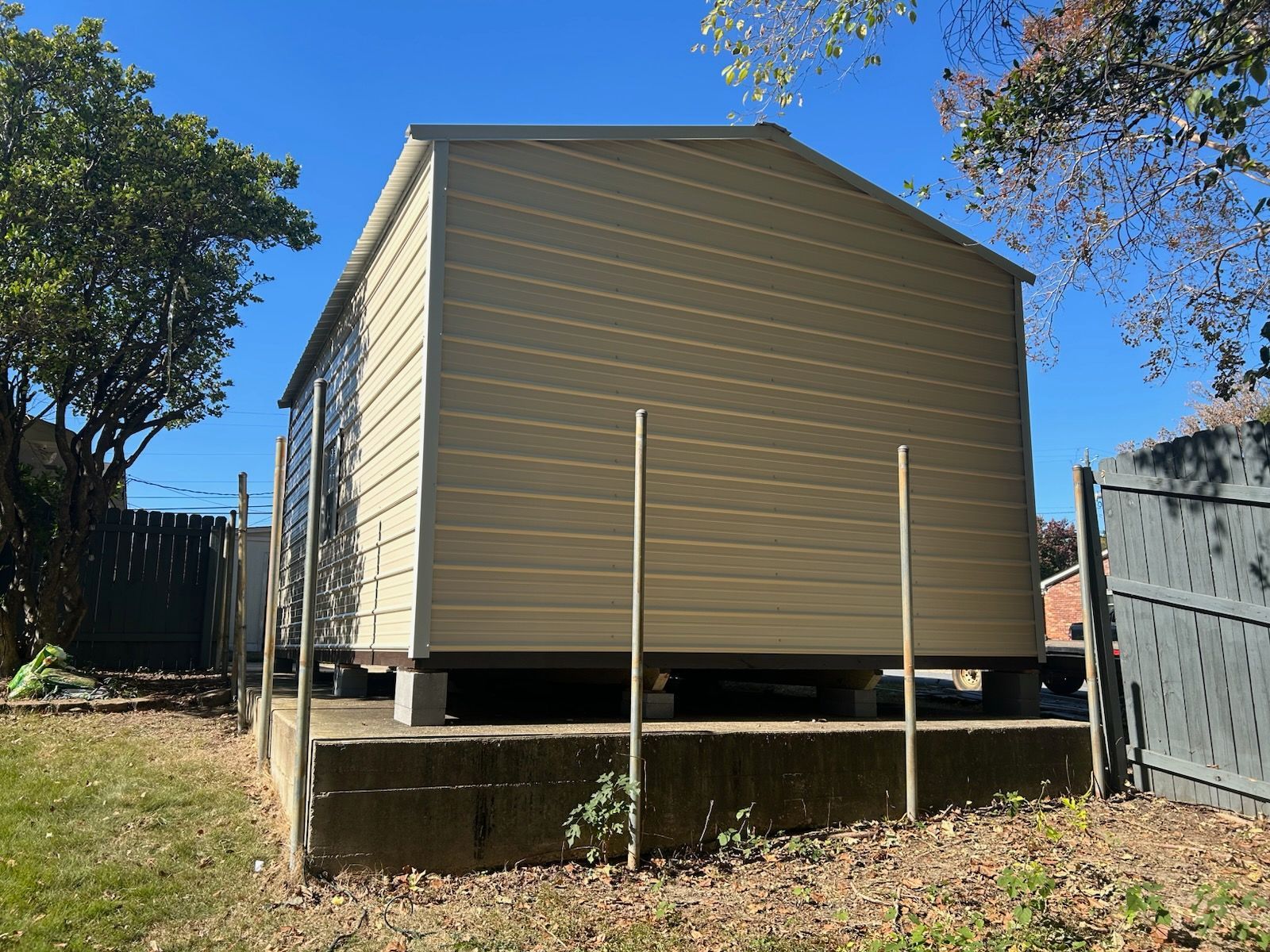 Tan shed on a concrete foundation with wooden supports. Backyard setting, blue sky, trees on each side.