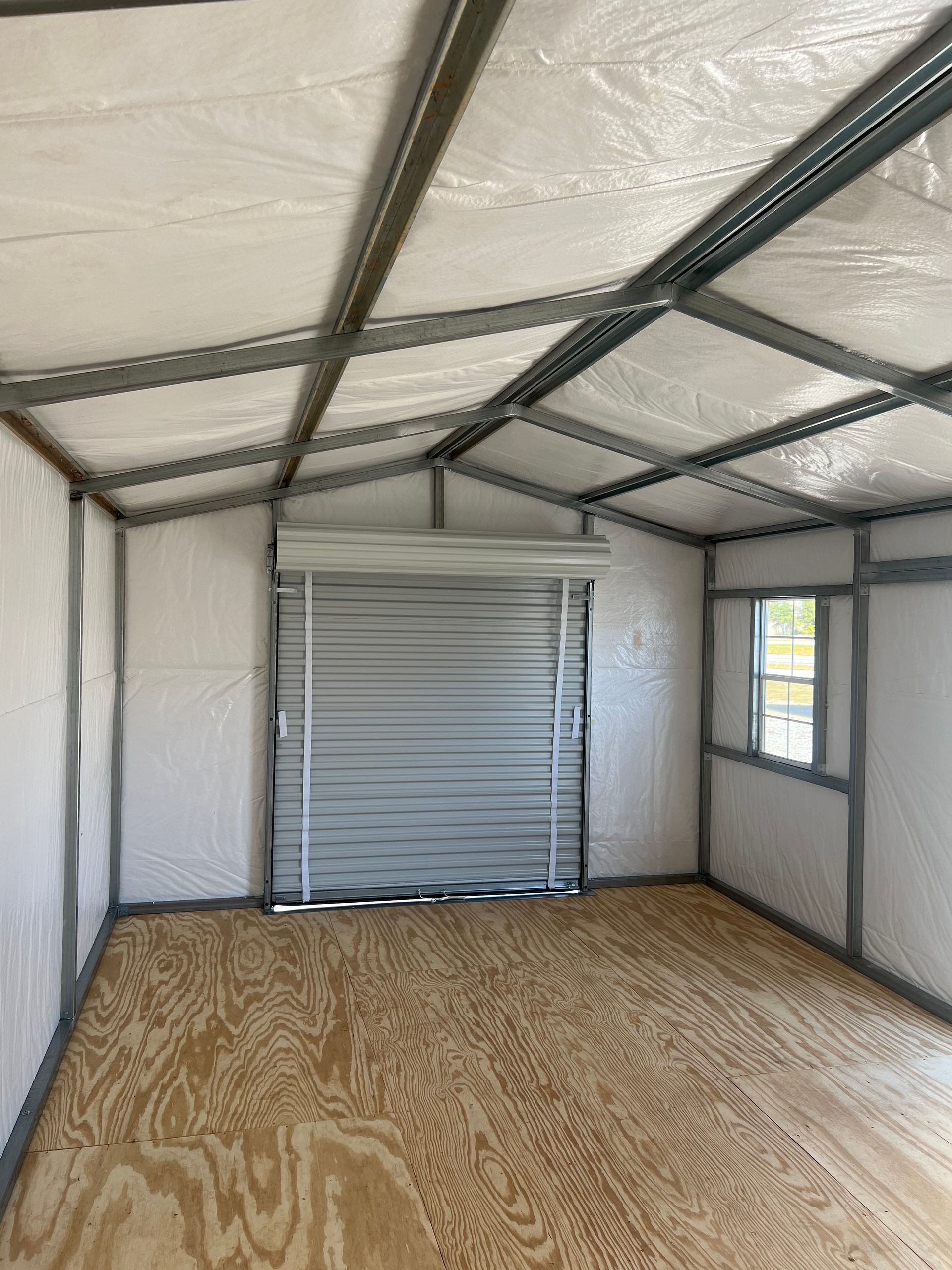 Interior of a shed with metal frame and roll-up door, featuring plywood floor and white walls.